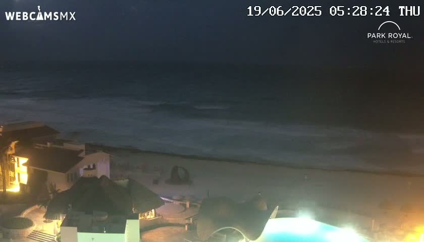 A nighttime view of a beach with moderately rough waves, illuminated buildings, and a partially visible swimming pool.