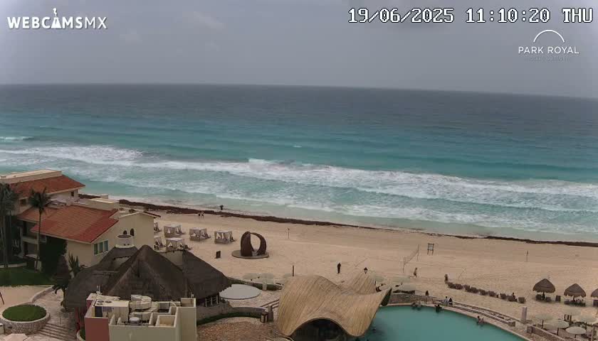 An overcast day shows a beach with turquoise water, moderate waves, and a resort with a pool overlooking the ocean.