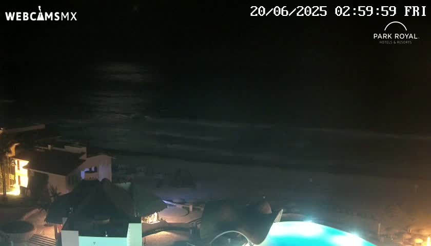 A nighttime view of a beach with calm waves, illuminated buildings, and a lit pool.