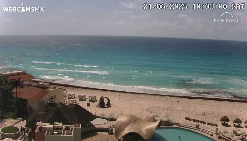 An aerial view shows a resort overlooking a beach with turquoise water and small waves under a partly sunny sky.