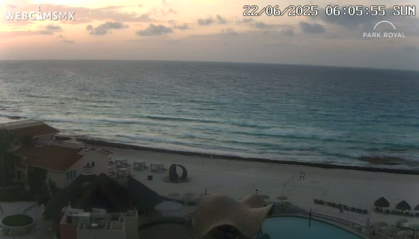 An ocean beach scene at sunrise shows calm waves lapping a sandy shore in front of a resort with a pool.