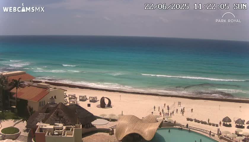 A beach with turquoise water and small waves under a partly cloudy sky, featuring a resort with buildings, a pool, and people enjoying the beach.