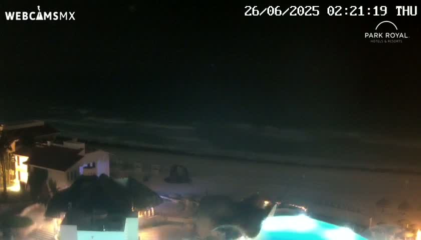 A nighttime view shows a beach with gentle waves under a clear, dark sky, overlooking illuminated buildings and a swimming pool.