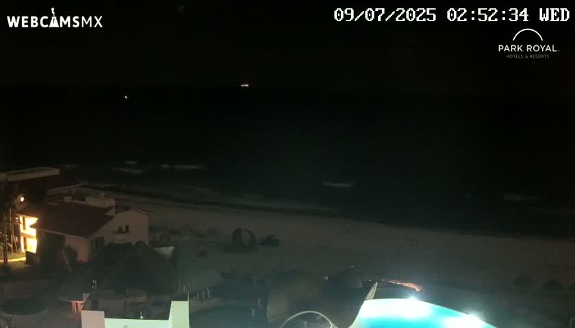 A nighttime view of a dark ocean with small waves, a sandy beach, and buildings with lights illuminated on the shore.