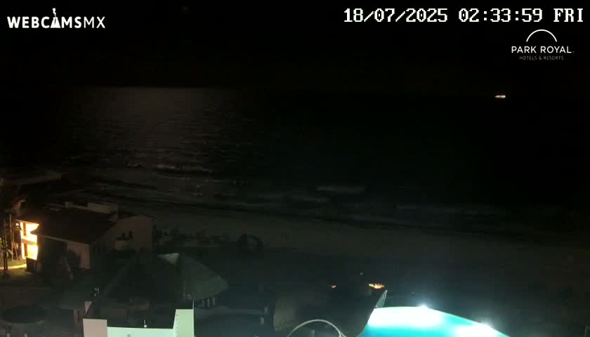 A nighttime view of a dark ocean, a sandy beach, and buildings with lights on, under a clear sky.