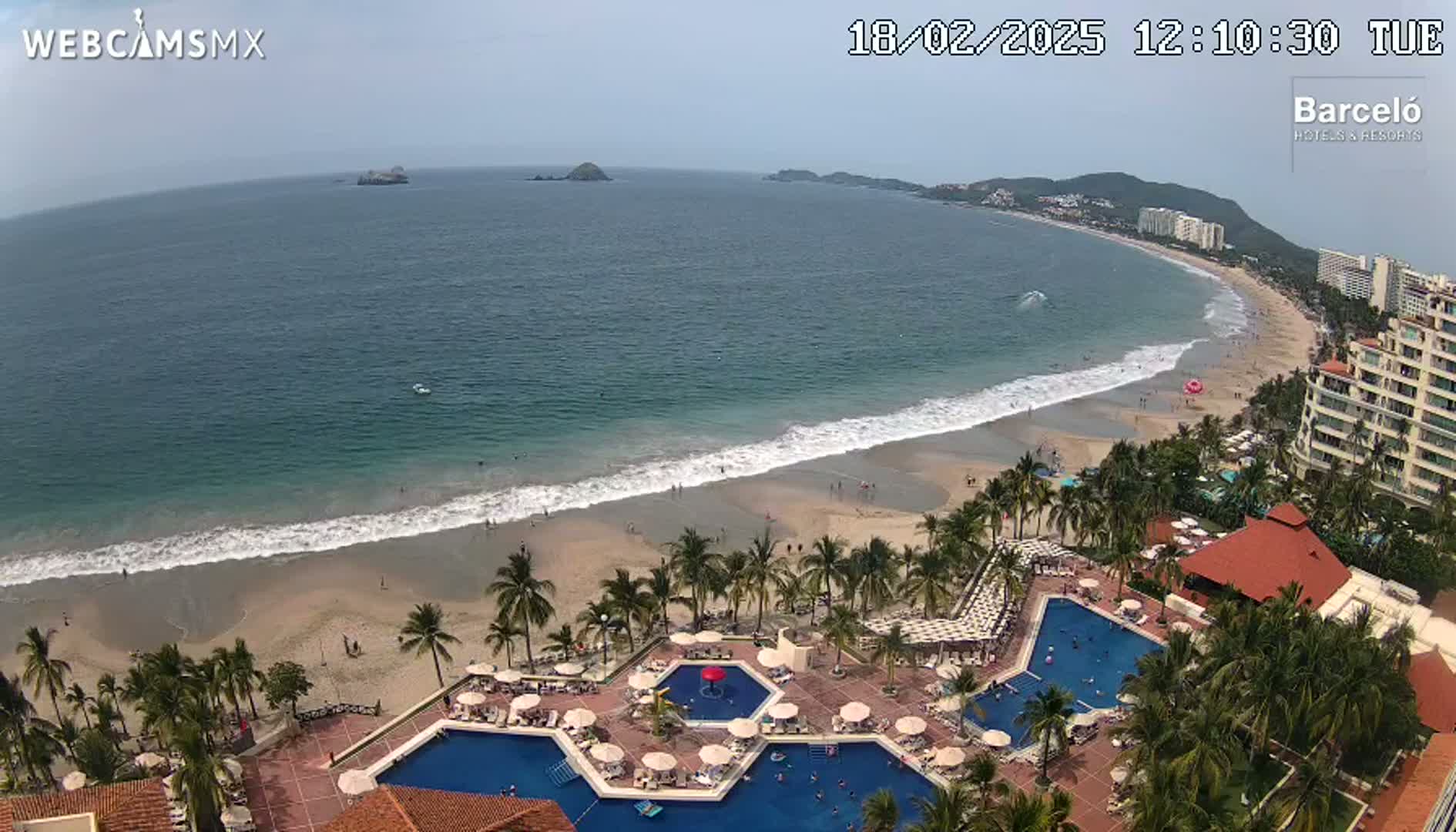 An aerial view shows a resort with multiple swimming pools next to a sandy beach with gentle waves on a partly cloudy day.
