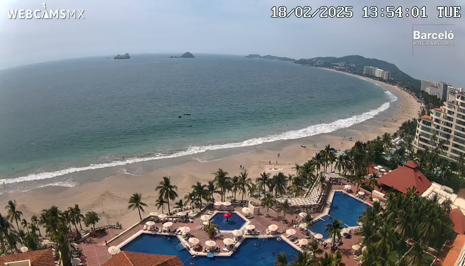An aerial view shows a resort with multiple swimming pools situated on a sandy beach next to a calm ocean under a mostly sunny sky.