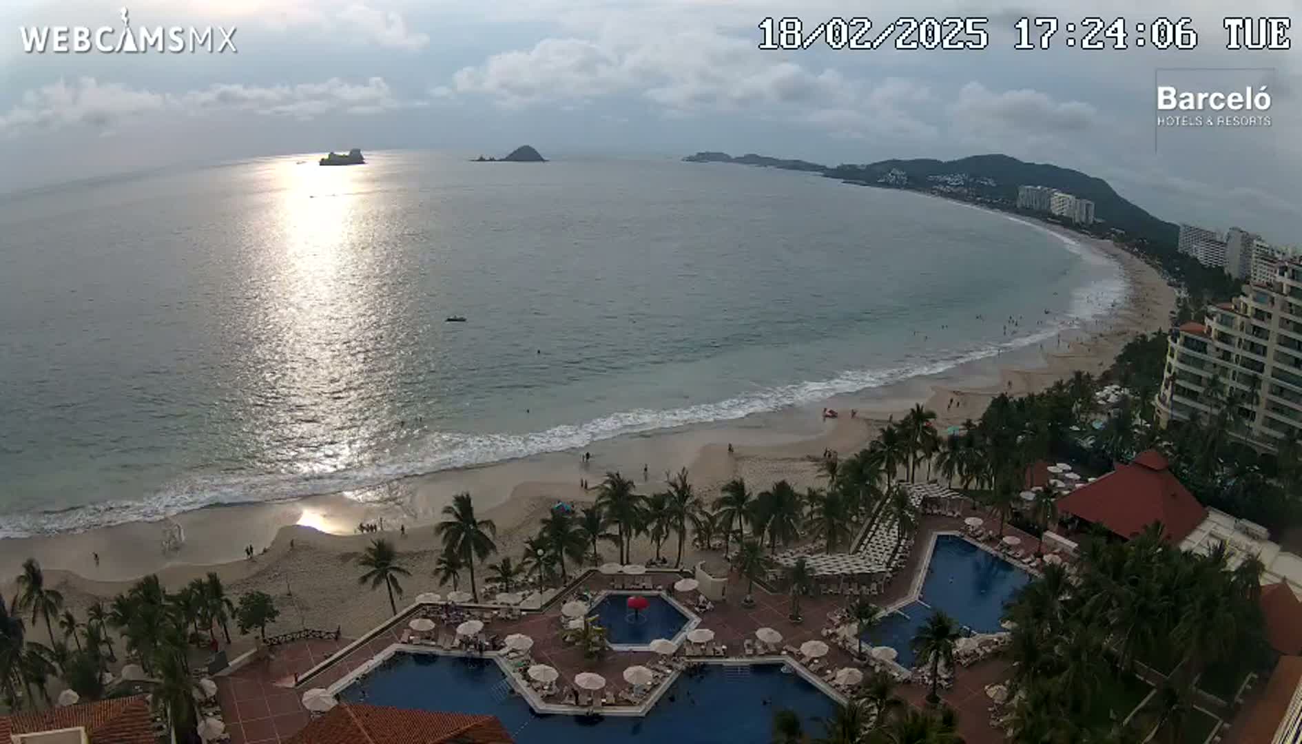 An ocean beach scene at sunset, with a resort featuring multiple pools, is viewed from above under partly cloudy skies.