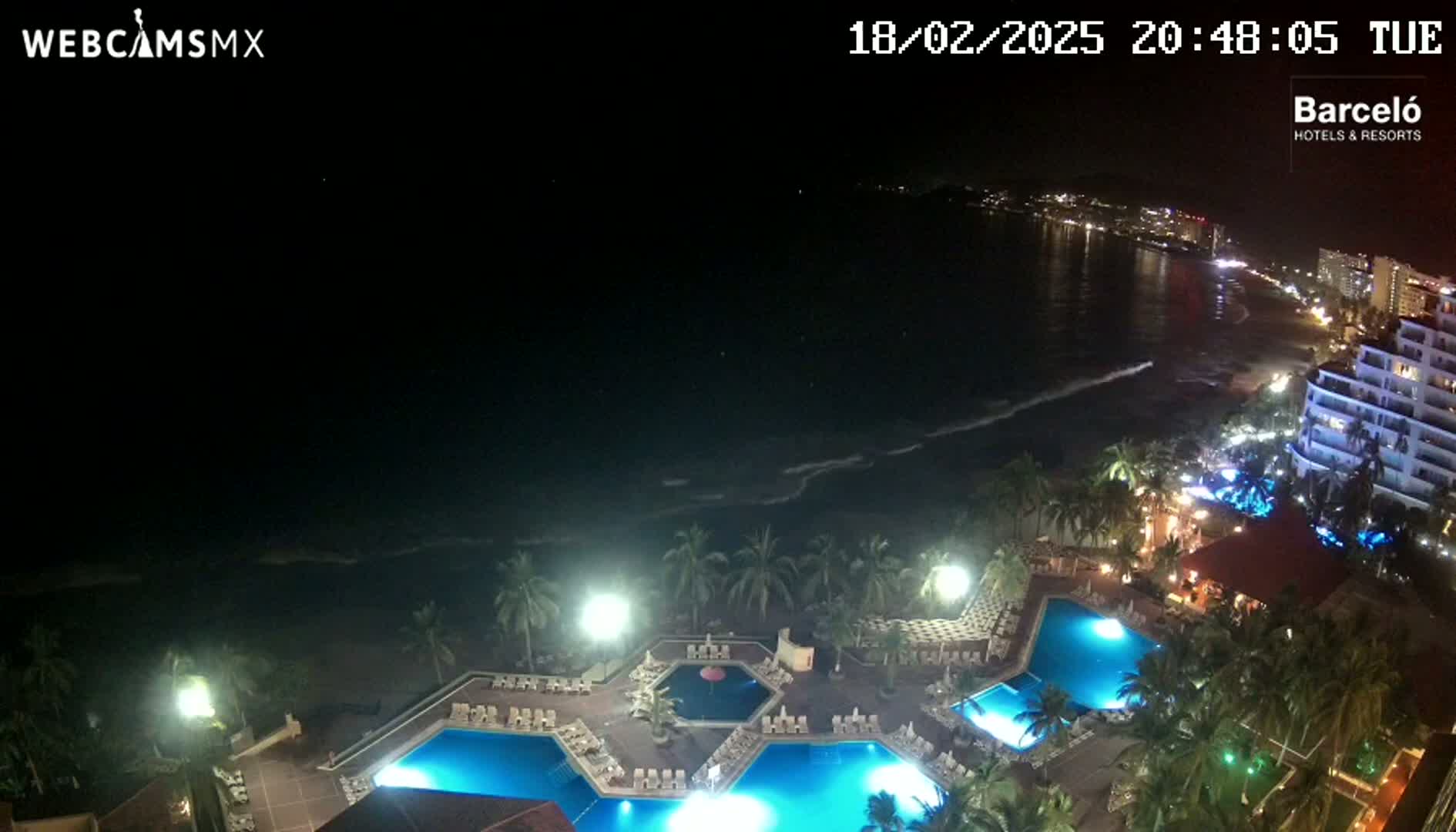 A nighttime aerial view shows a resort with illuminated pools and palm trees overlooking a dark ocean with gentle waves and a city skyline in the distance.