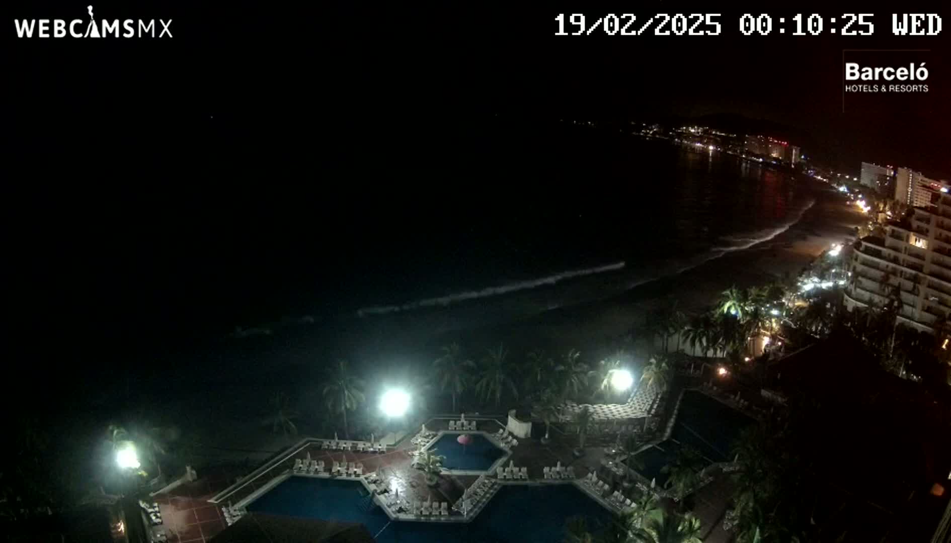 A nighttime view of a resort pool area with palm trees, a beach, and ocean waves under a clear, dark sky.