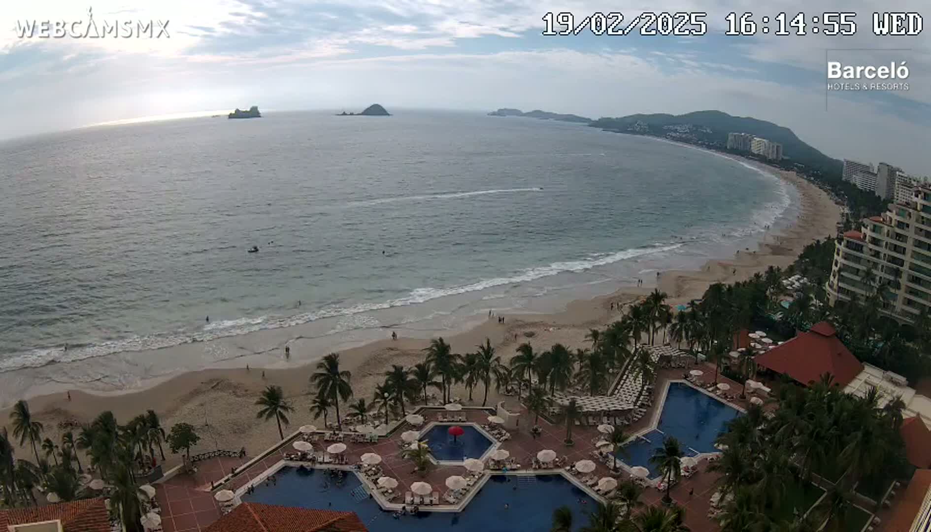 An aerial view of a resort with multiple swimming pools overlooking a sandy beach and calm ocean under a partly cloudy sky.