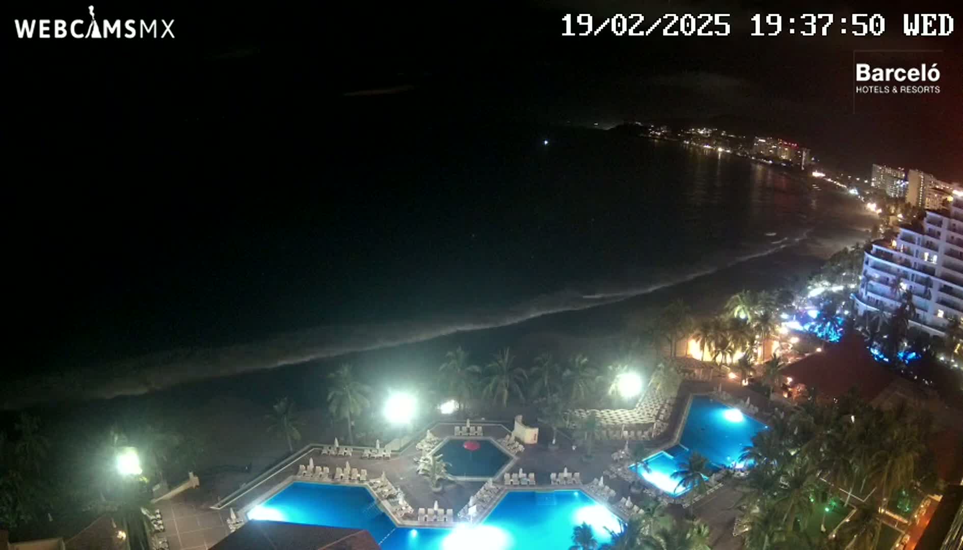 A nighttime, aerial view shows a resort with multiple illuminated pools next to a dark ocean with a gently lapping shoreline, under a clear, dark sky.
