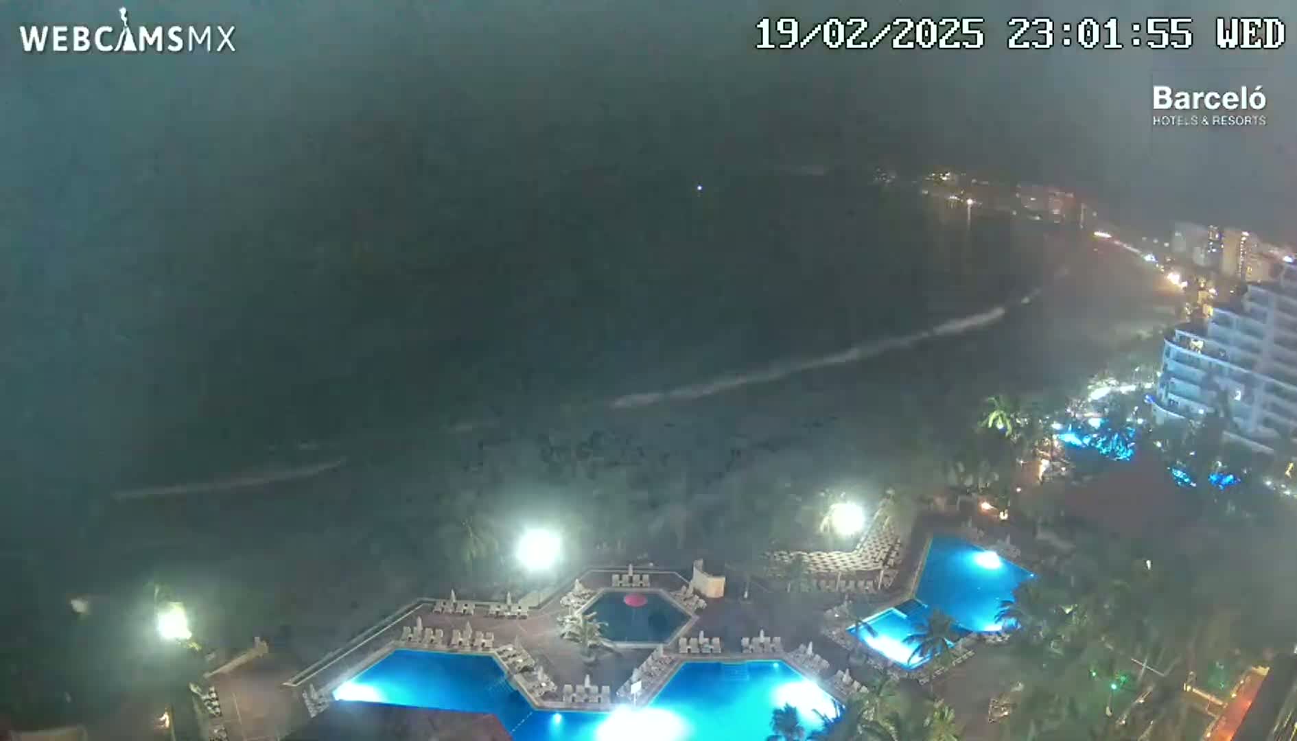 A nighttime aerial view of a resort with multiple illuminated pools, overlooking a stormy ocean with heavy rain.