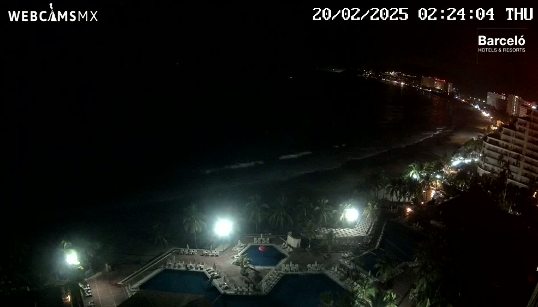 A nighttime view of a resort with multiple pools, illuminated by lights, overlooking a dark ocean with gentle waves and a dimly lit coastline.