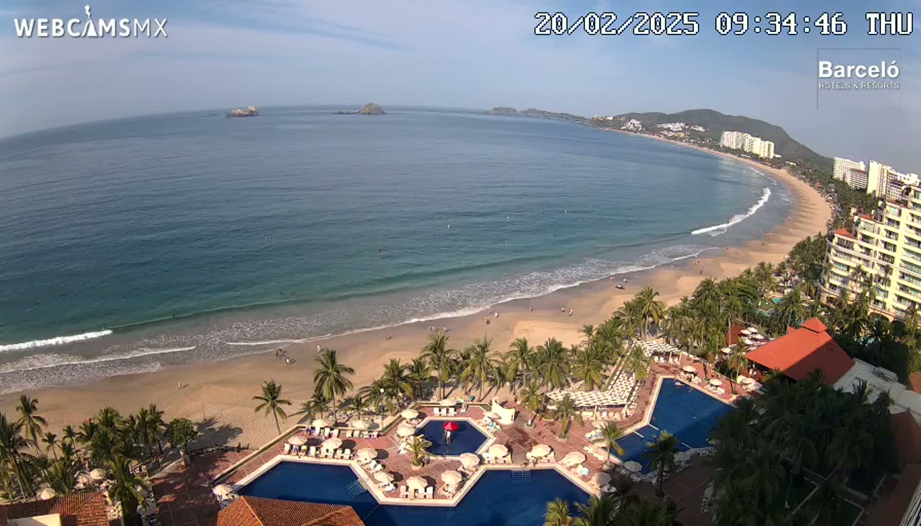 An aerial view shows a resort with multiple pools adjacent to a sandy beach and ocean under a mostly sunny sky.