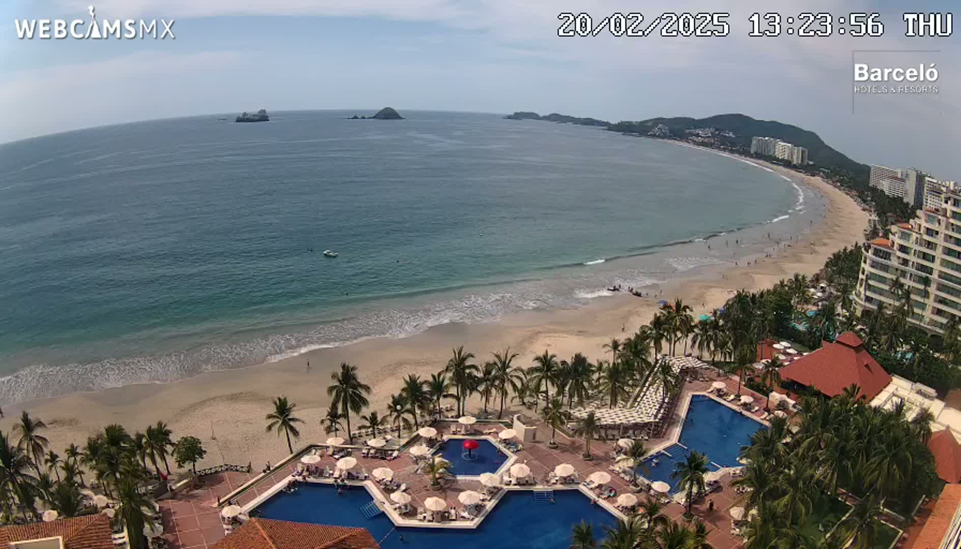 An aerial view shows a resort with multiple pools situated on a sandy beach next to a calm ocean under a mostly clear sky.