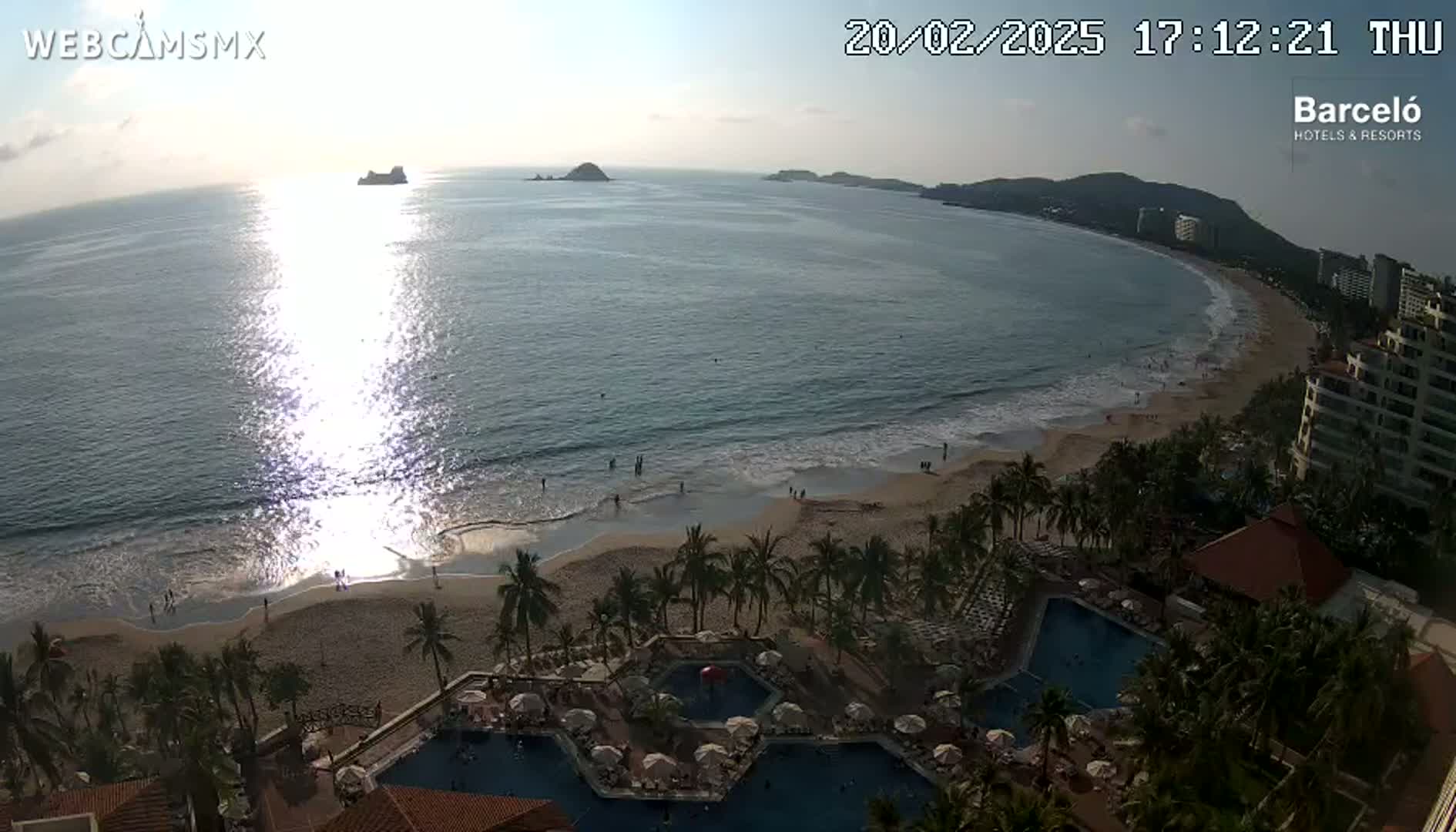 A beach with calm waves, palm trees, resort pools, and buildings under a mostly clear sunset sky.