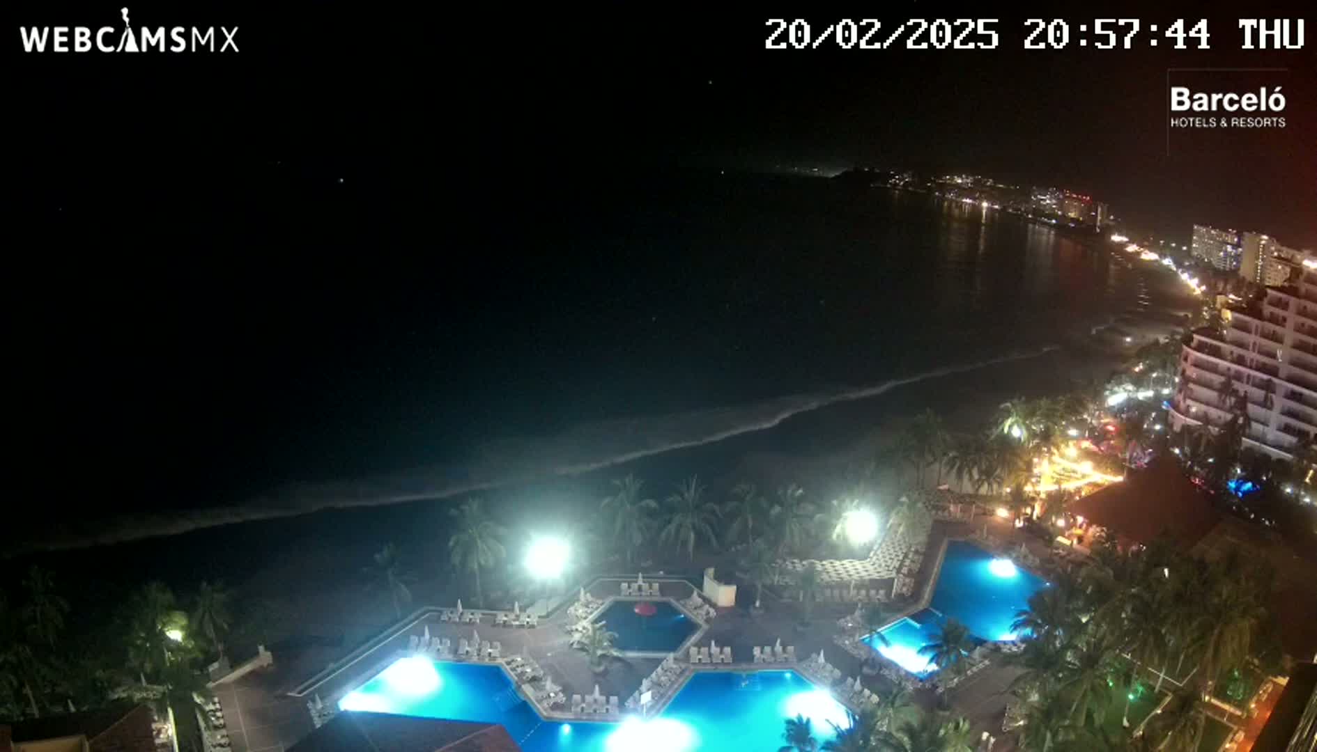 An illuminated resort with multiple pools overlooks a dark, ocean shoreline at night.