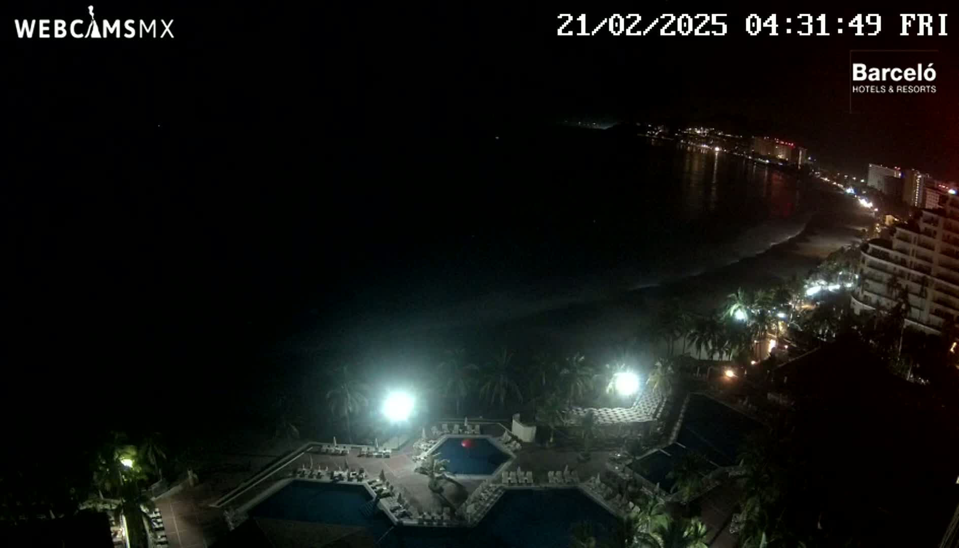 A nighttime view of a resort with multiple pools, surrounded by palm trees, overlooking a dark ocean and a city skyline under a clear sky.