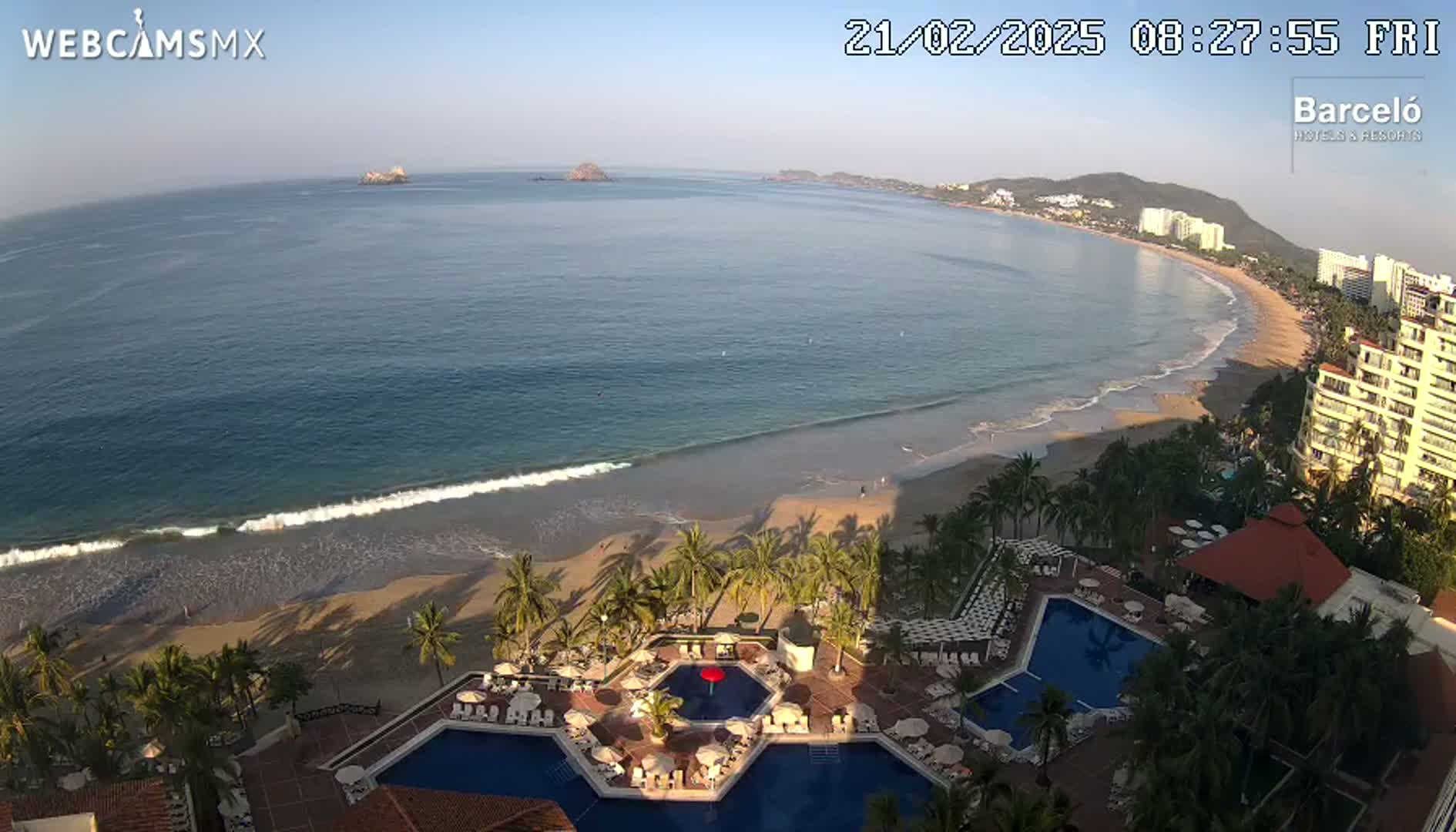 An aerial view shows a resort with multiple pools overlooking a sandy beach and calm ocean under a clear sky.