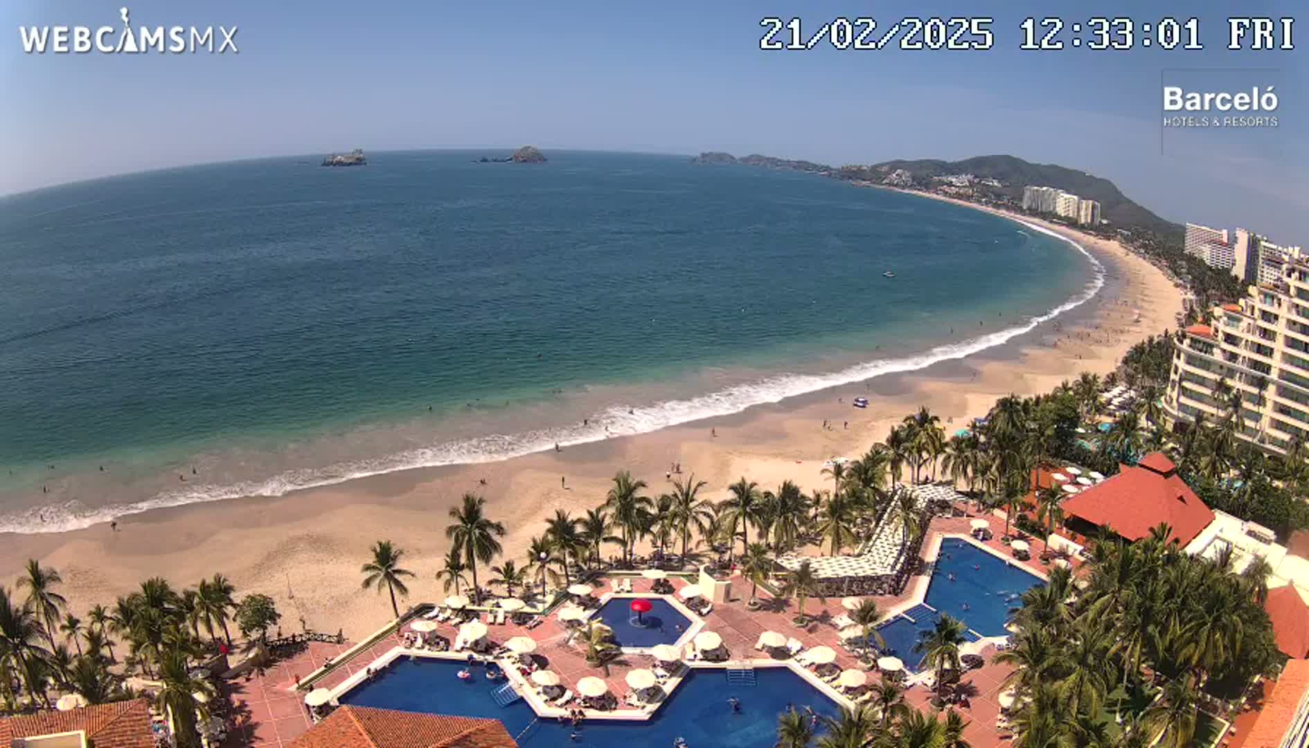 An aerial view shows a long sandy beach with calm ocean waves under a clear blue sky, alongside a resort with multiple swimming pools surrounded by palm trees.