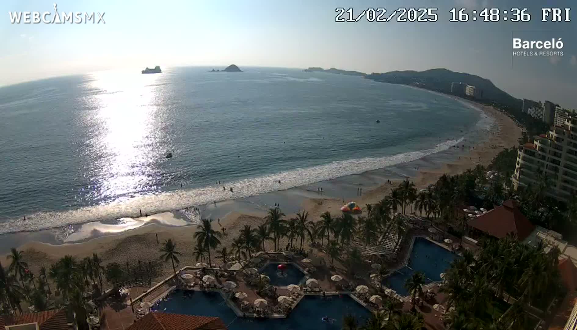 An aerial view of a beach resort on a sunny day, showing a sandy beach, ocean waves, palm trees, and multiple swimming pools.