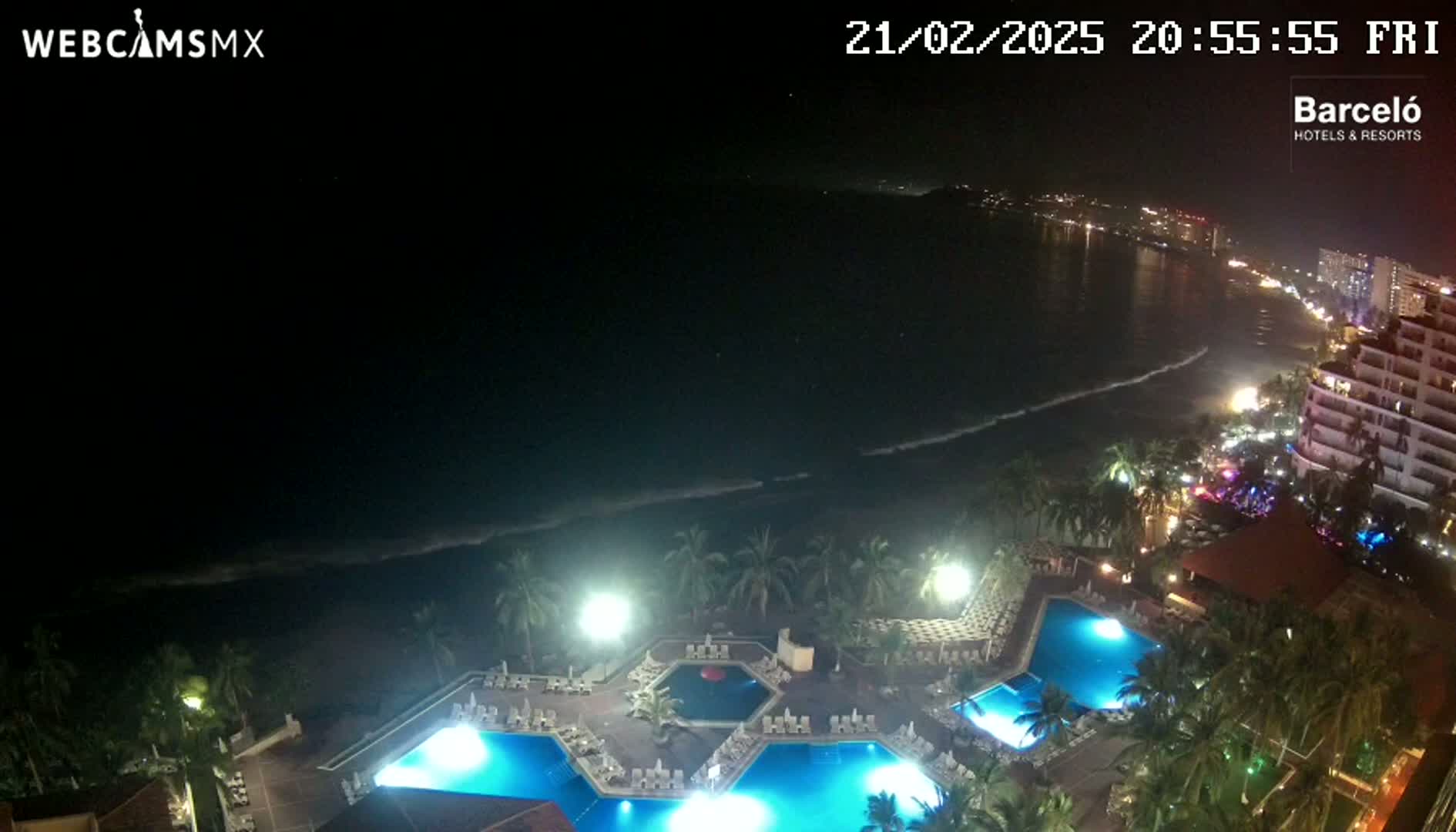 A nighttime aerial view of a resort complex with multiple illuminated pools, palm trees, and a beach with gently lapping waves under a dark, clear sky.