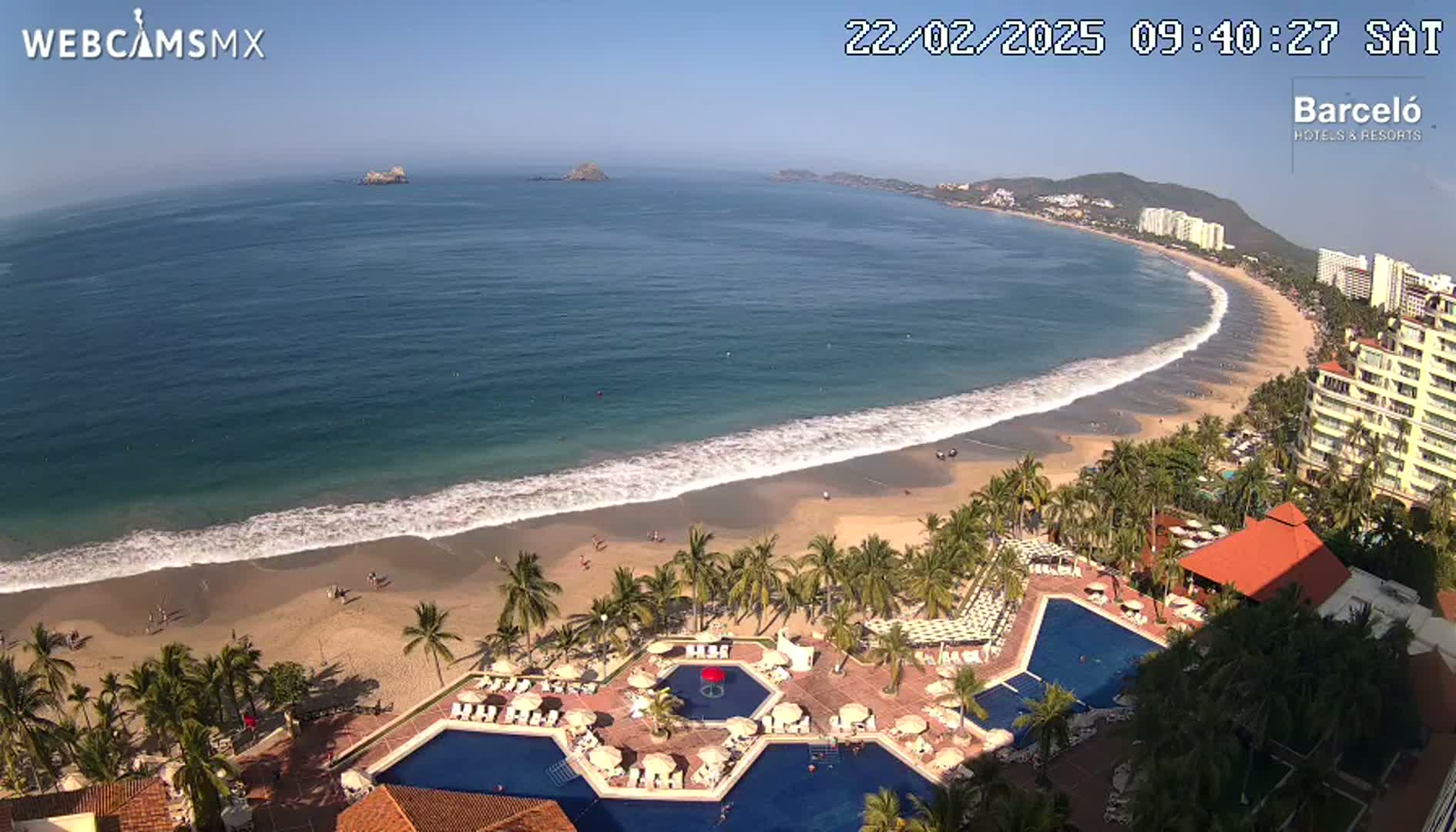 An aerial view shows a long, sandy beach with gentle waves lapping the shore under a clear, sunny sky, adjacent to a resort with multiple pools surrounded by palm trees.