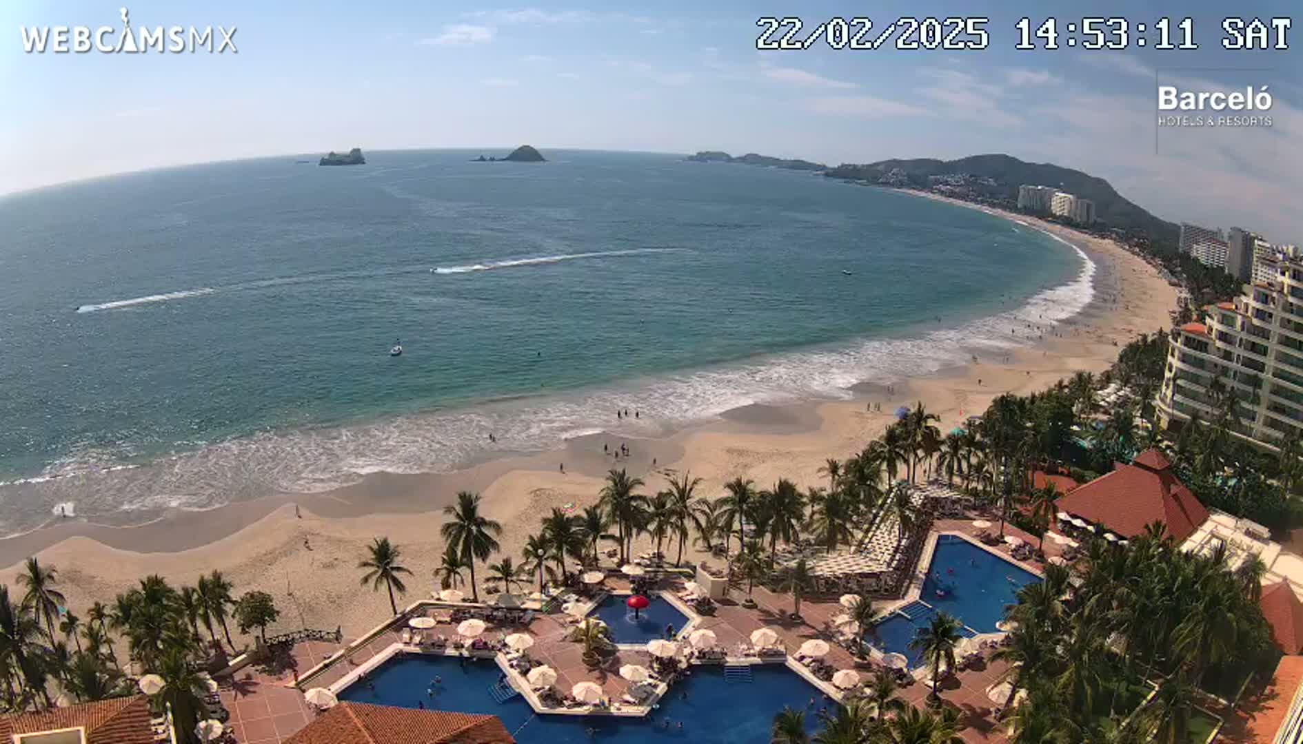 An aerial view shows a resort with multiple swimming pools nestled amongst palm trees next to a sandy beach and a calm ocean under a sunny sky.