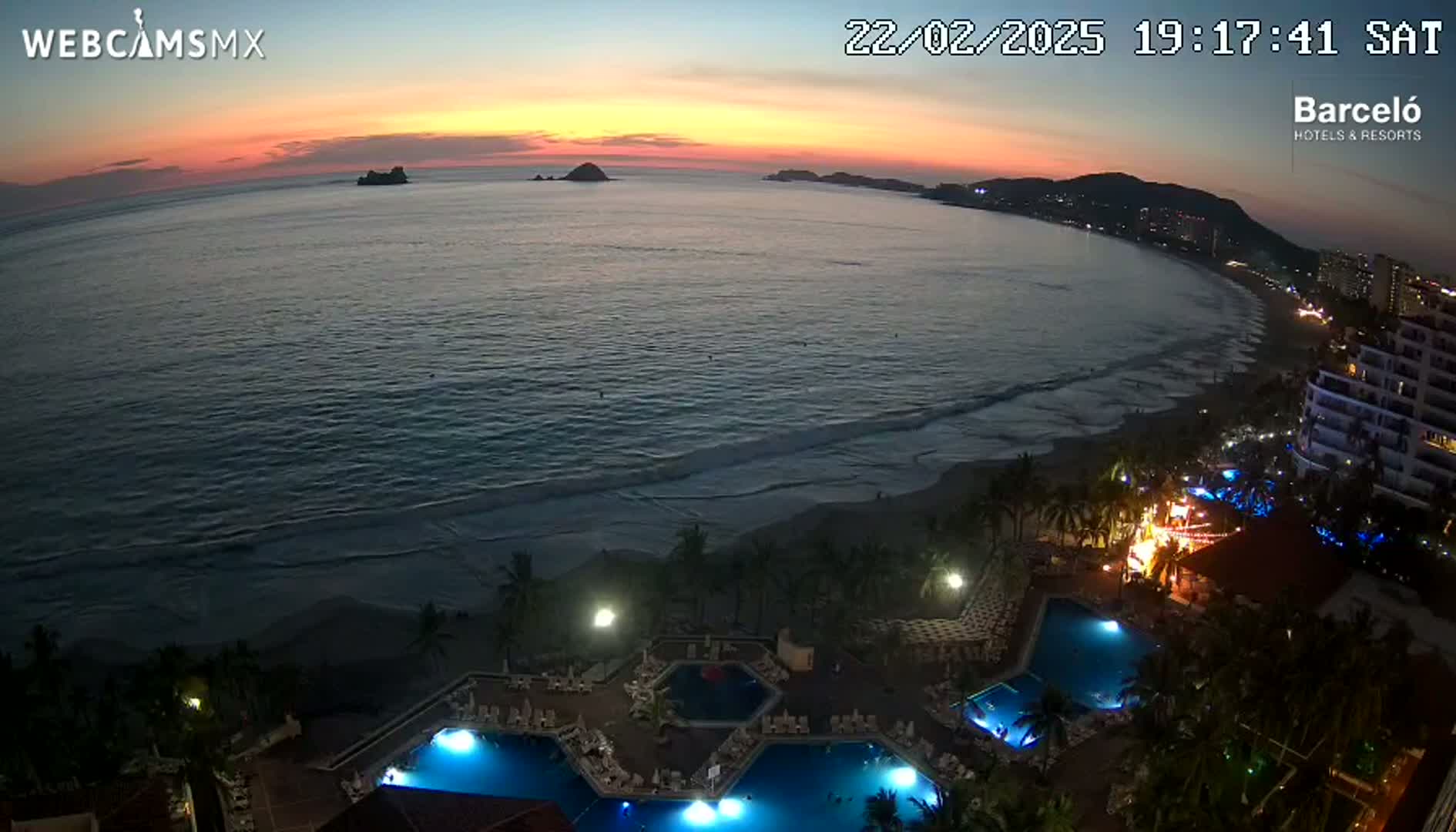 An ocean beach at sunset is viewed from above, showing a resort with illuminated pools and a shoreline with gently rolling waves.