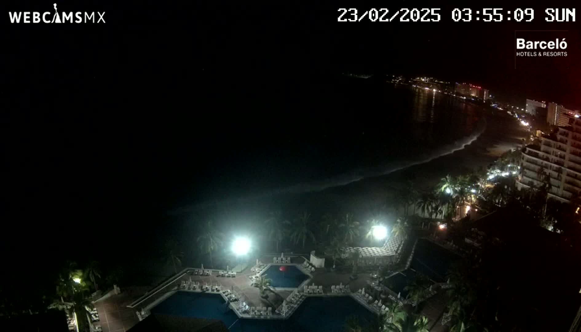 A nighttime view from a high vantage point shows a resort with multiple illuminated pools, palm trees, and a dark ocean with waves lapping a beach lined with lights.