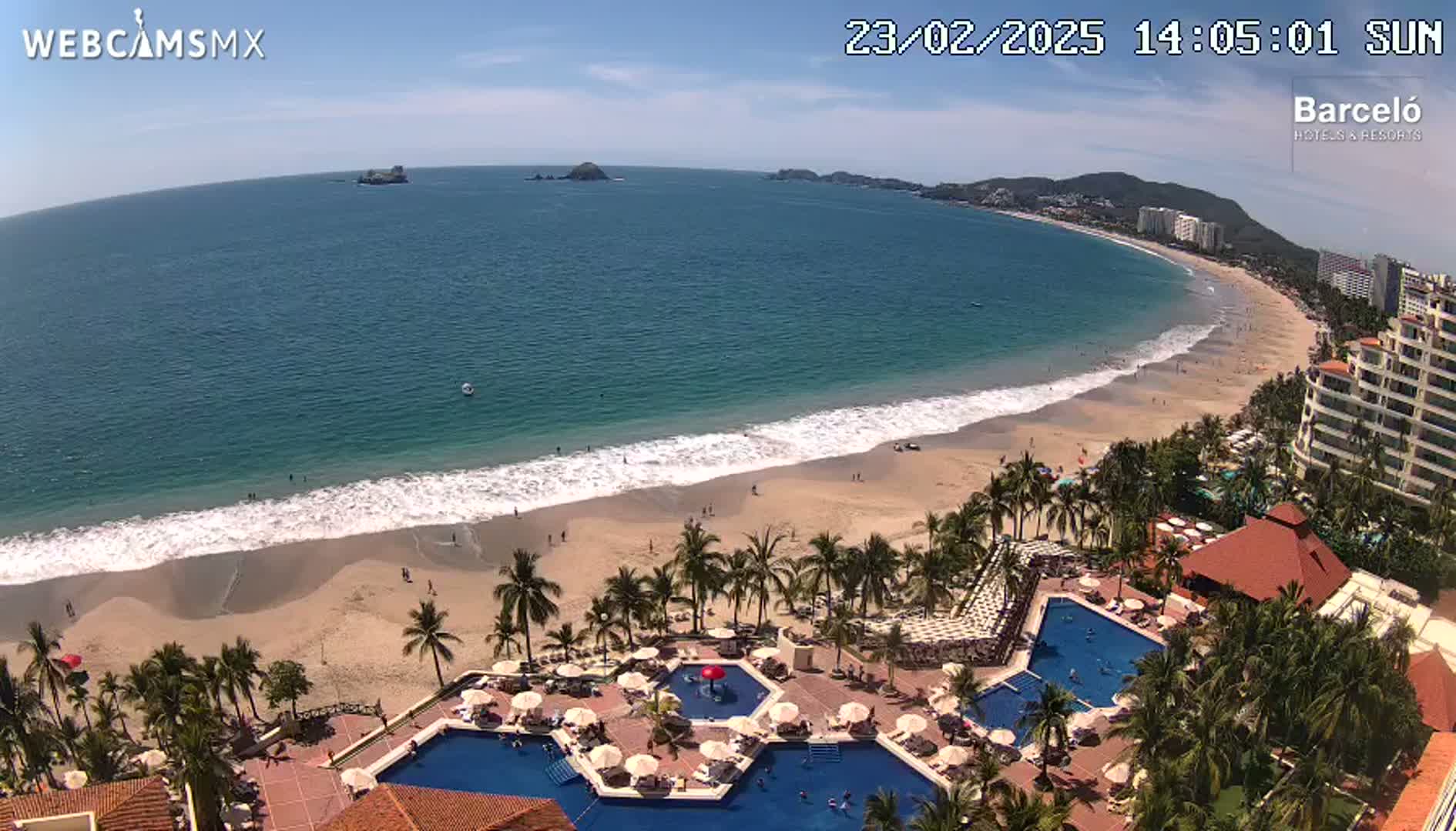 An aerial view shows a beach with calm waves, palm trees, and a resort with multiple pools under a sunny sky.