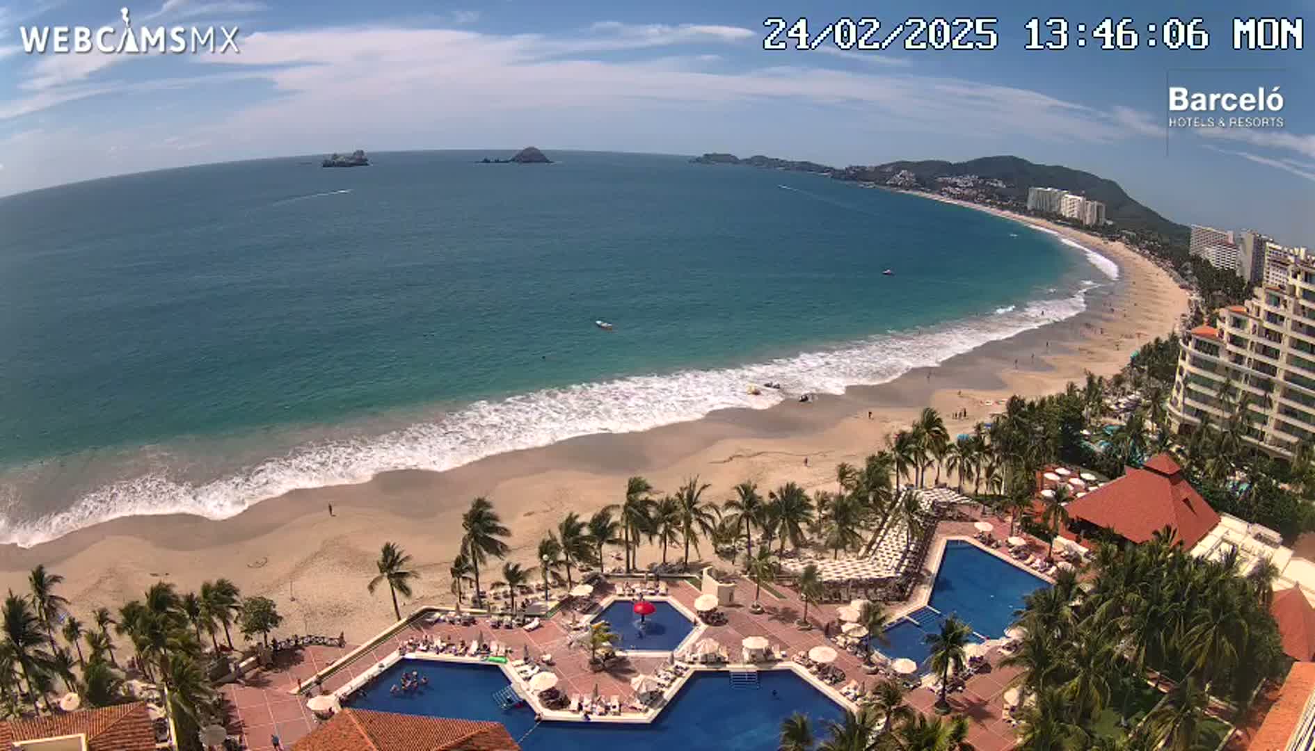 An aerial view shows a resort with multiple pools overlooking a sandy beach and calm, blue ocean under a partly cloudy sky.