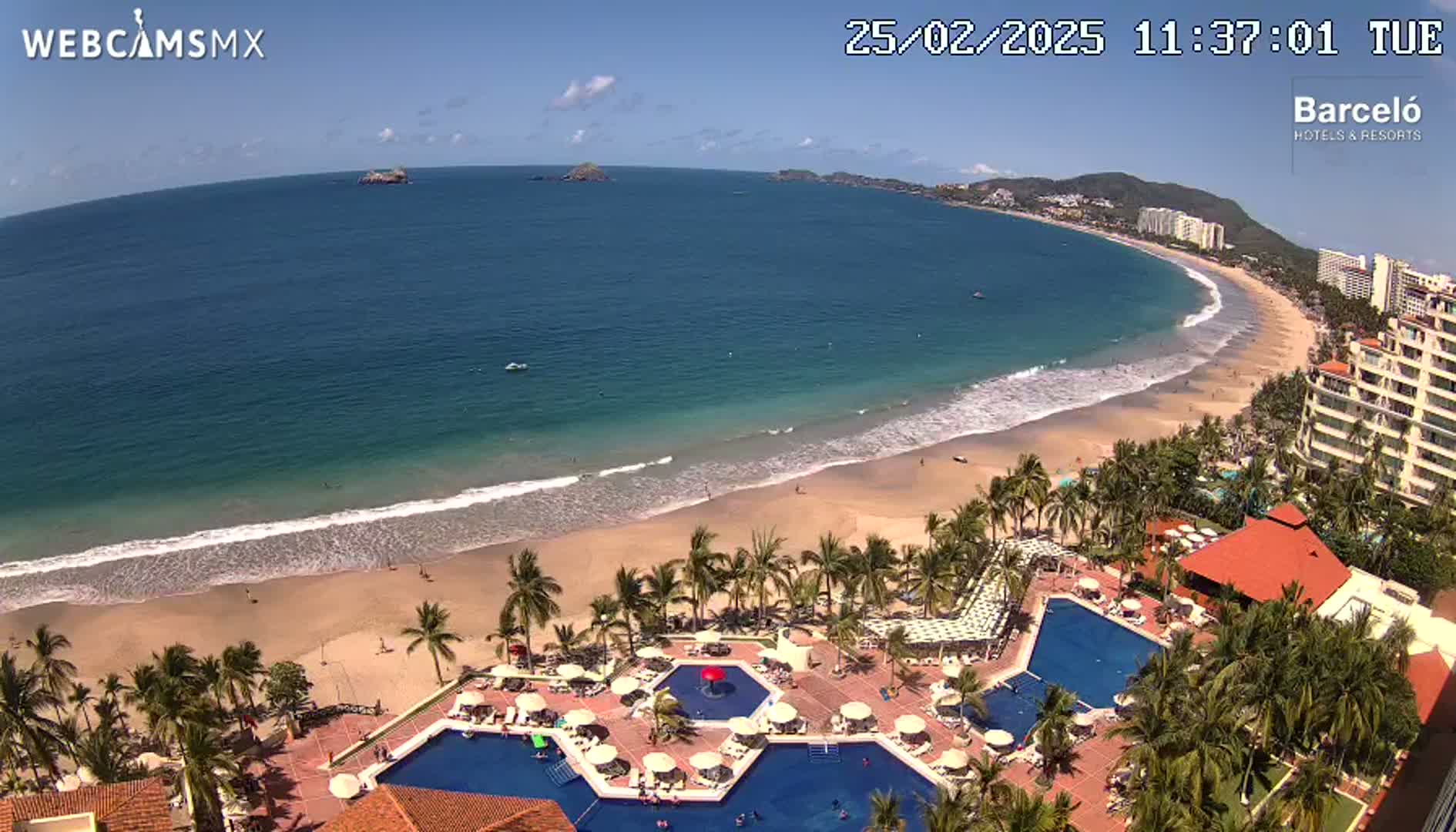 A sunny beach with gentle waves laps a sandy shore lined with palm trees and a resort featuring multiple pools.