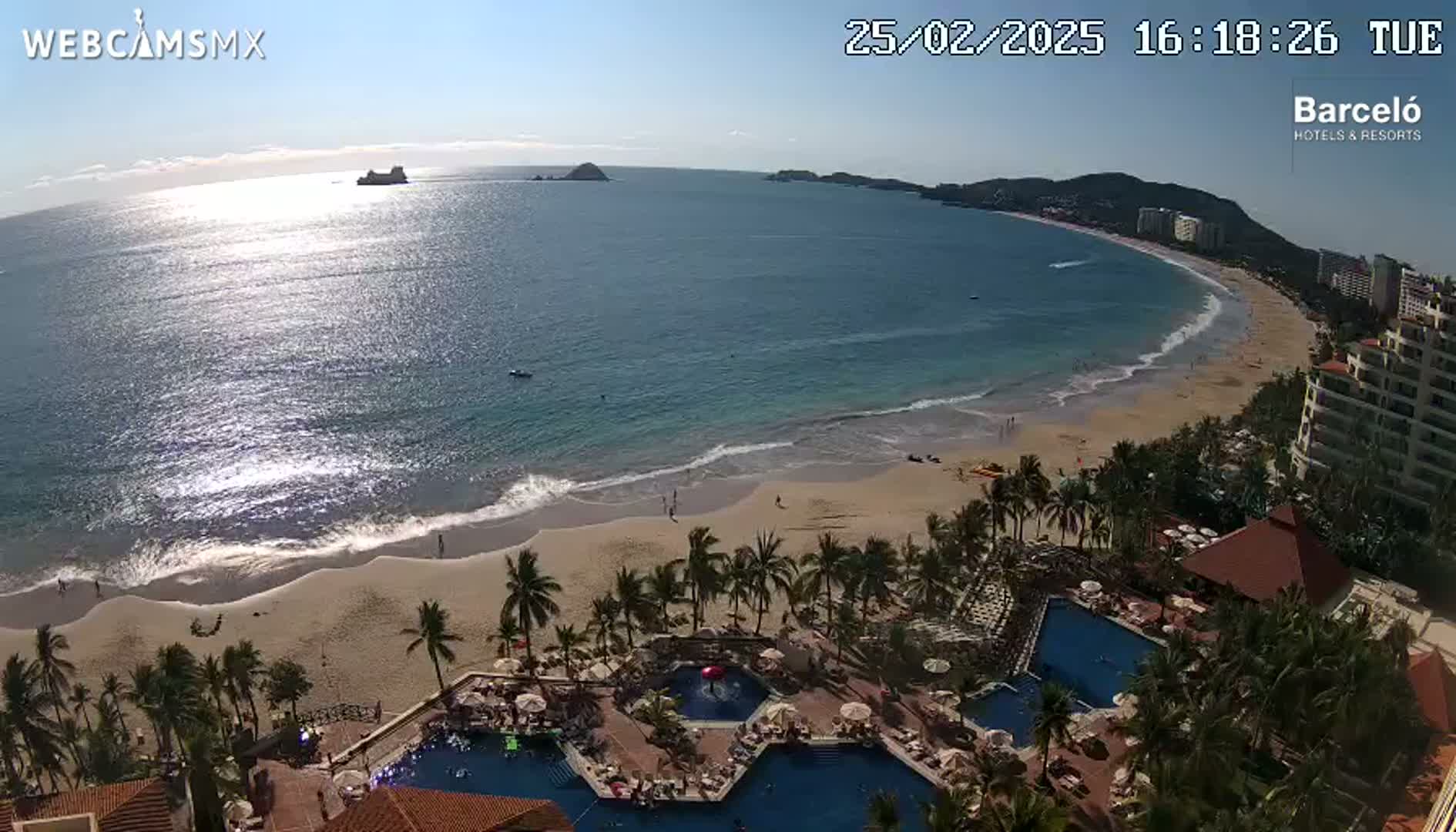 An aerial view shows a beach with calm waves under a sunny sky, overlooking a resort with multiple pools surrounded by palm trees.