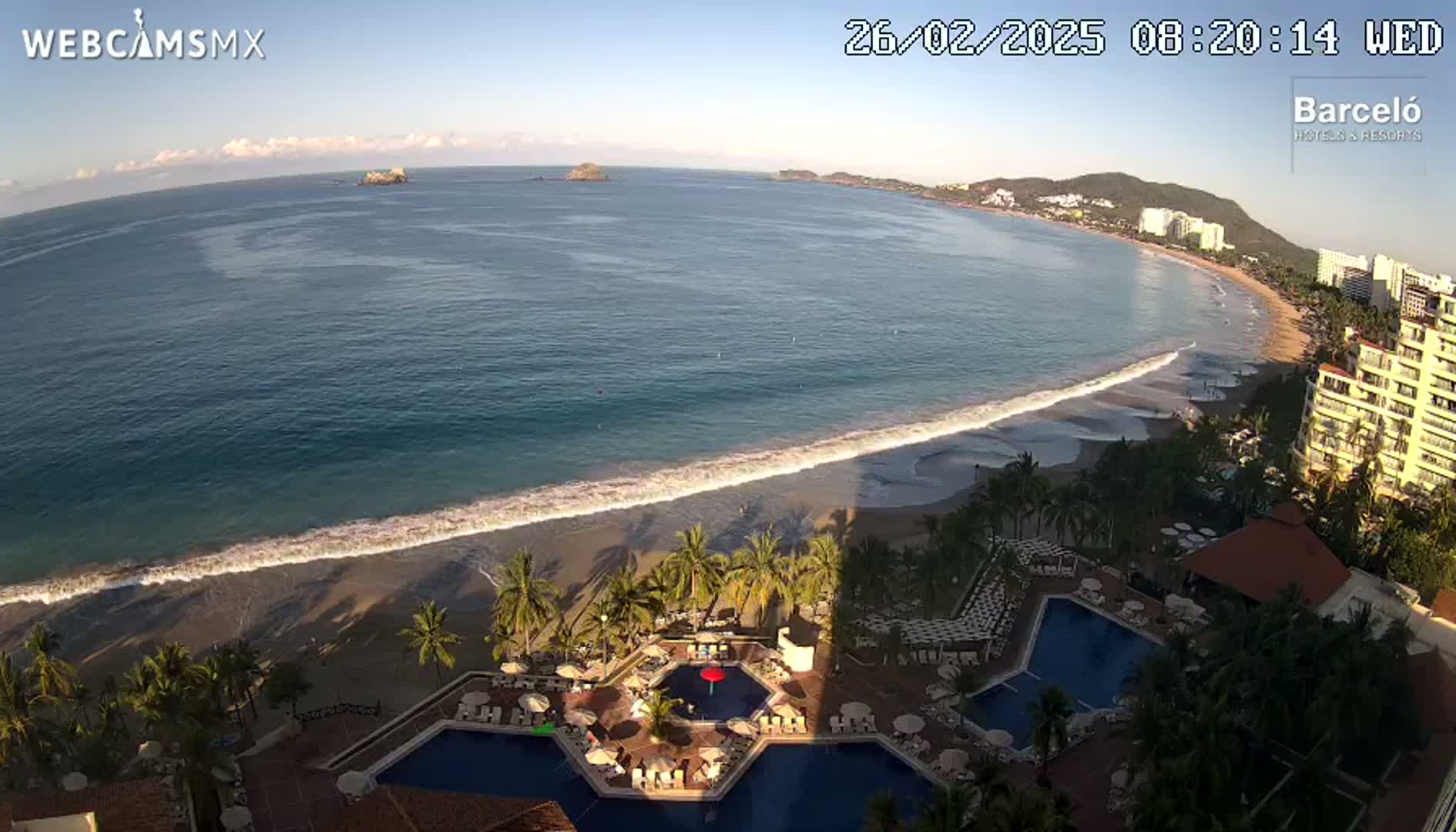 An aerial view shows a beach with gentle waves lapping the shore, palm trees, hotel pools, and buildings under a clear, sunny sky.