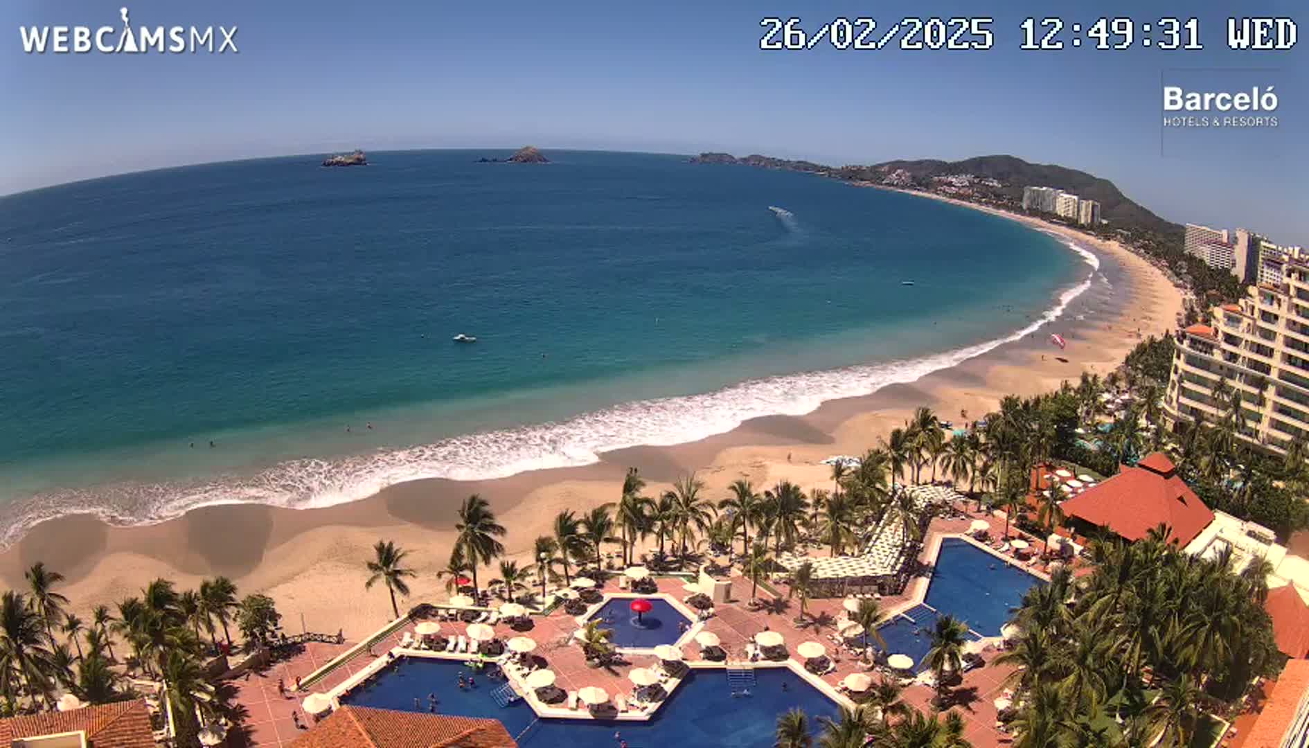 An aerial view of a beach resort on a sunny day, showing a long sandy beach with gentle waves lapping the shore, palm trees lining the beach, and multiple swimming pools within the resort grounds.