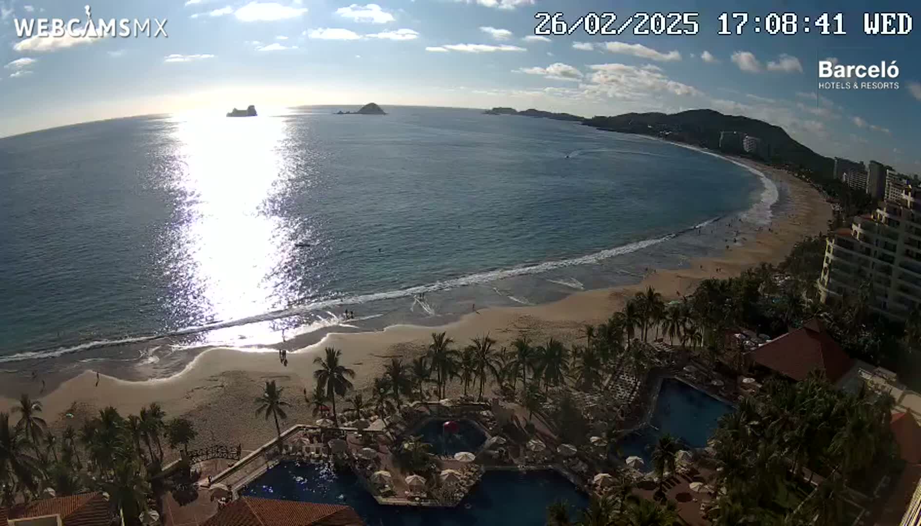 A beach with calm waves and a resort with pools is seen at sunset, under partly cloudy skies.