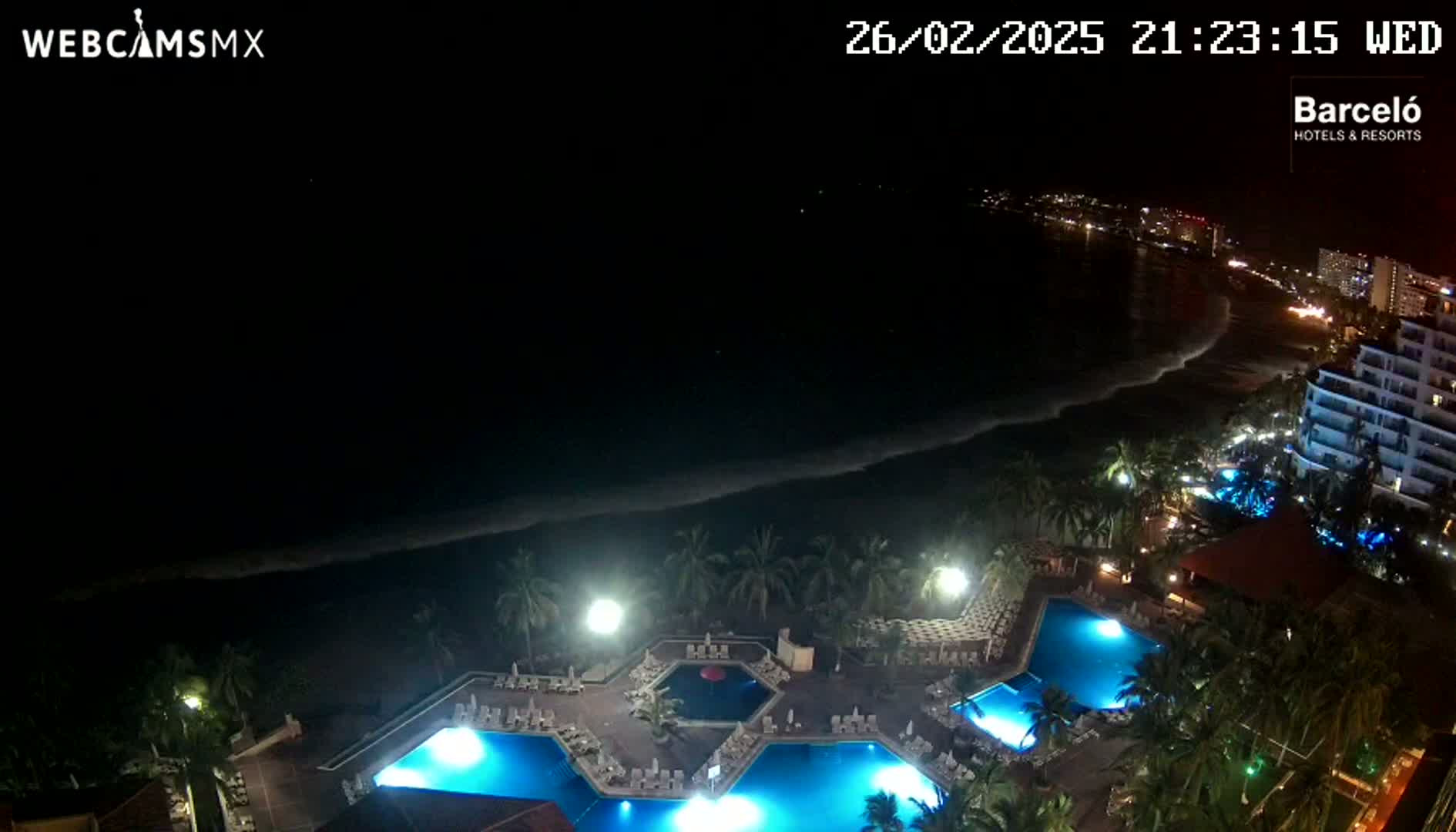 A nighttime aerial view shows a resort with illuminated pools next to a dark ocean with waves under a clear, starless sky.