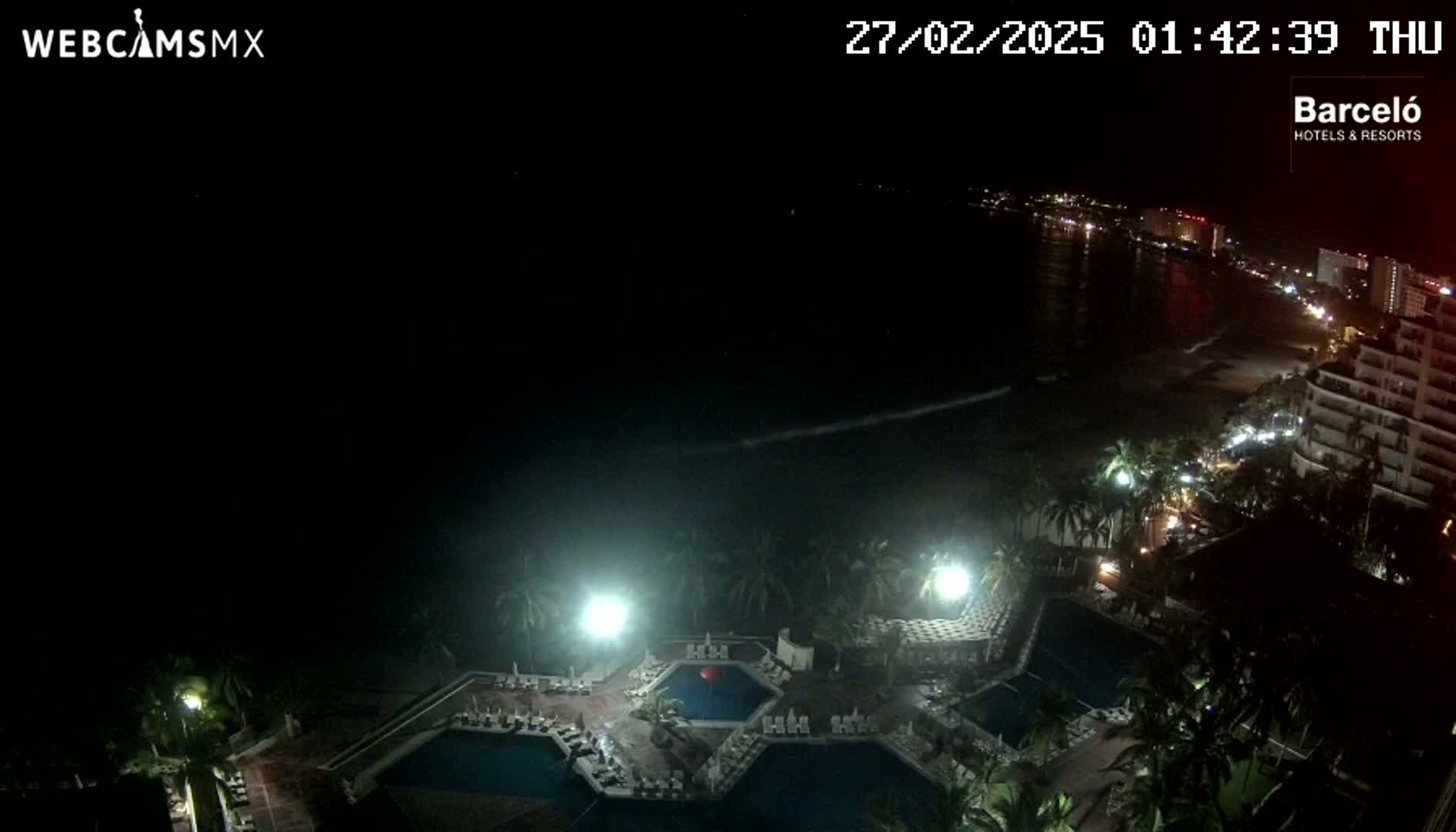 An aerial nighttime view shows a resort with multiple pools illuminated, palm trees, and a dark ocean with a distant, faintly lit coastline.