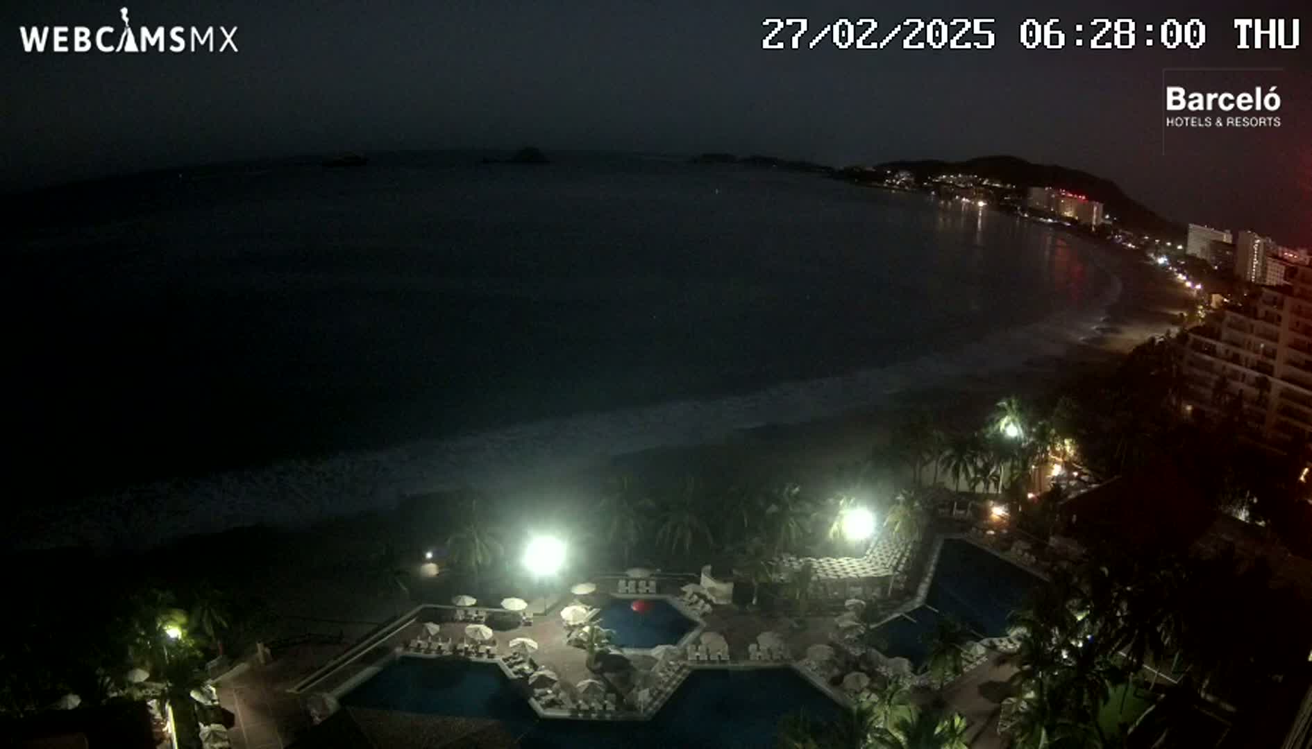 A nighttime aerial view shows a resort with illuminated pools and palm trees next to a dark ocean with gentle waves under a dark sky.