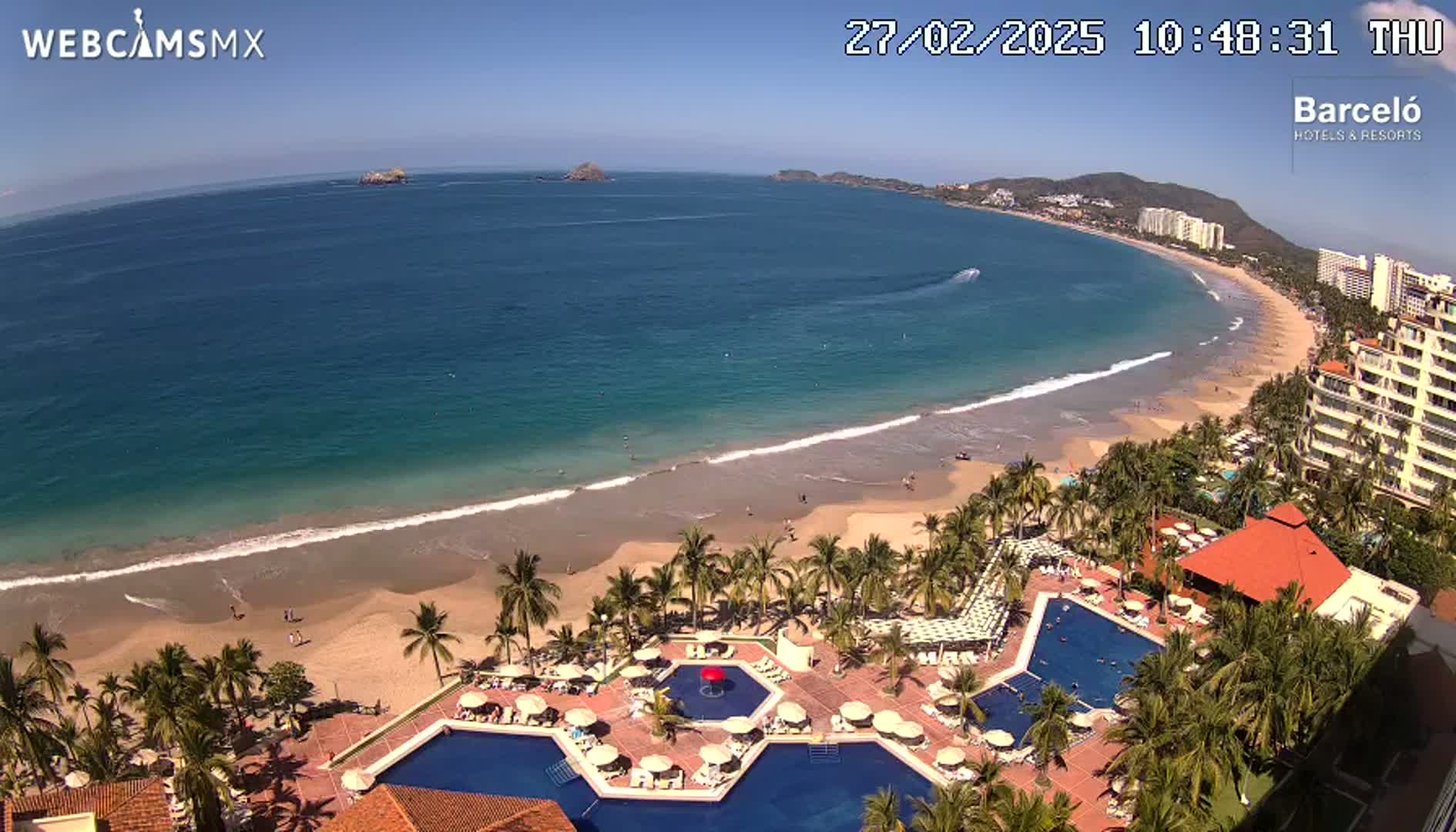 An aerial view shows a resort with multiple pools next to a sandy beach and calm ocean under a clear blue sky.