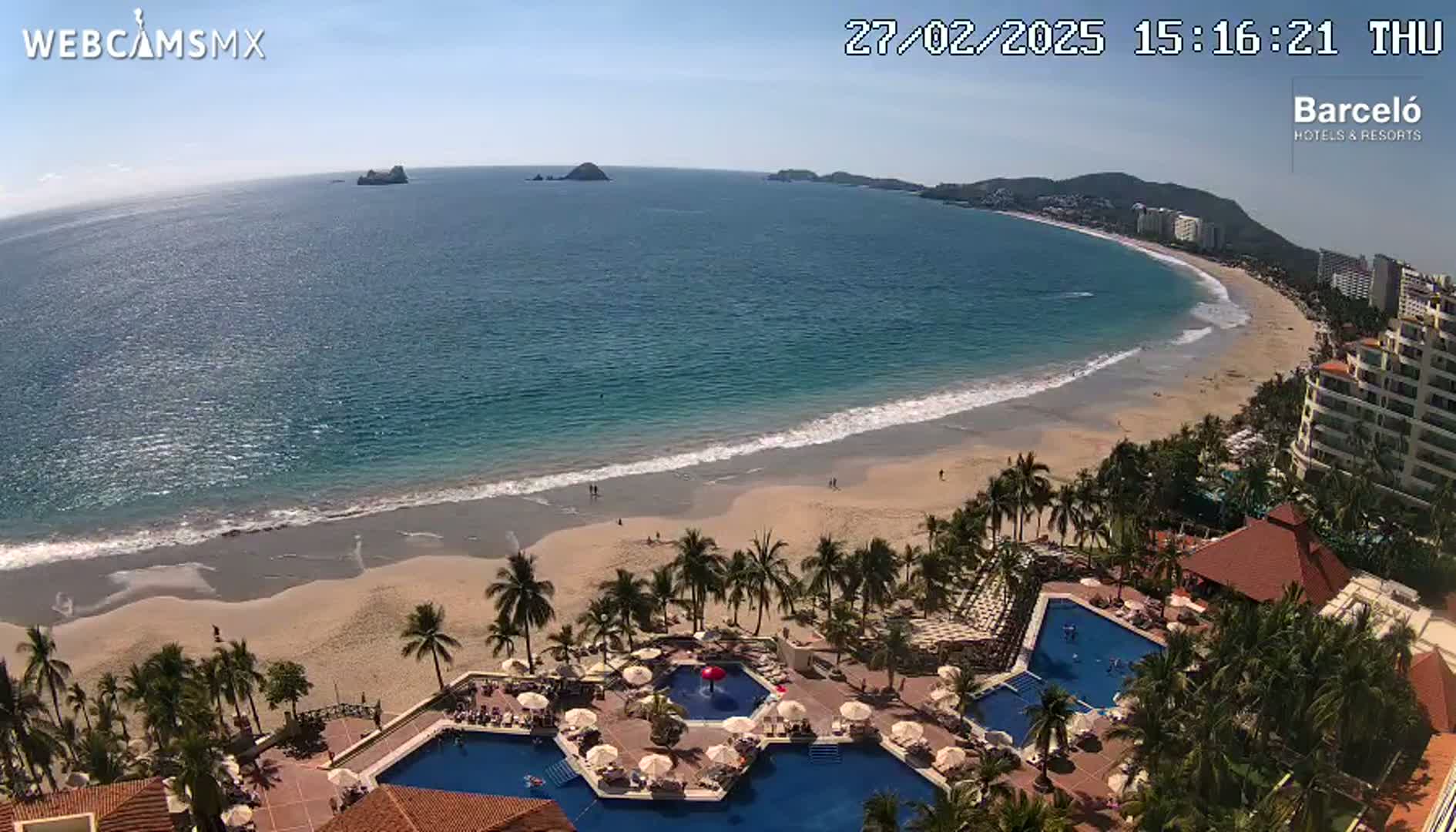 An aerial view shows a beach with calm ocean waves under a sunny sky, and a resort with multiple pools surrounded by palm trees.