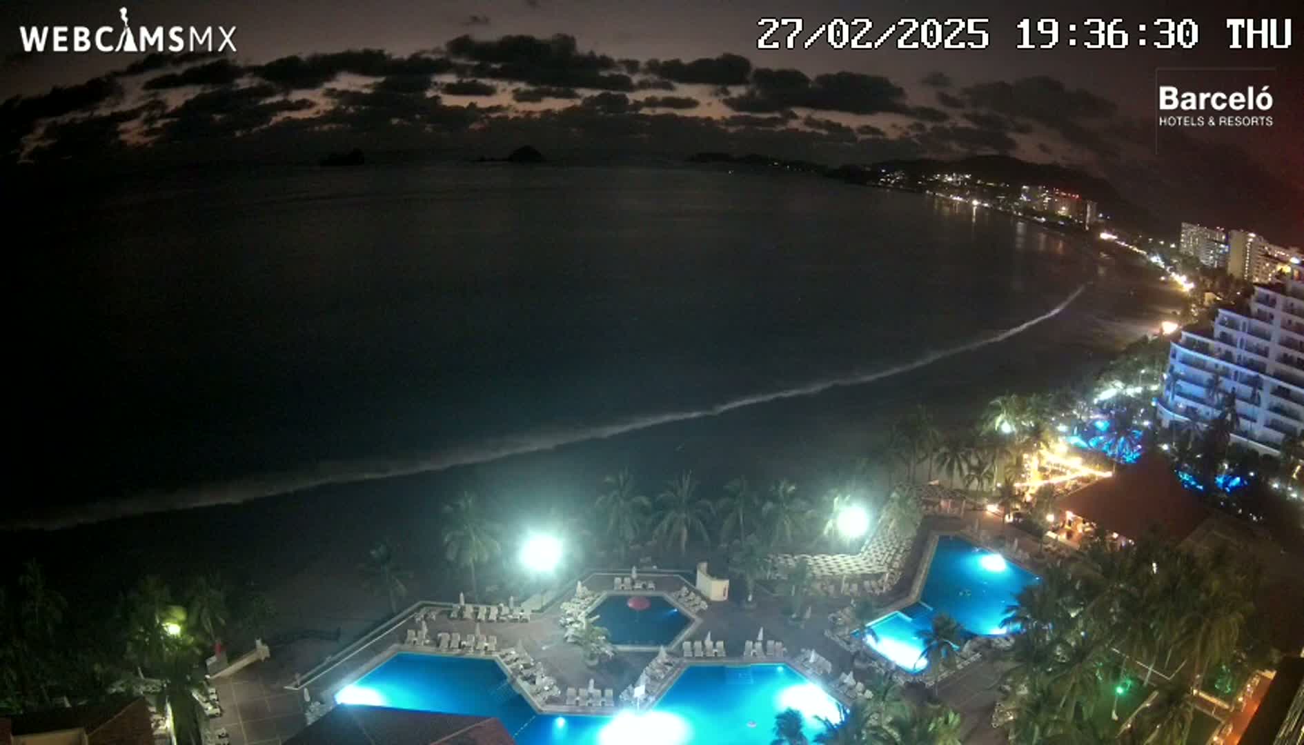 An illuminated resort with multiple pools overlooks a dark ocean with gentle waves under a partly cloudy night sky.