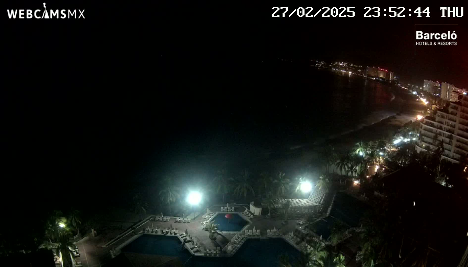 A nighttime, high-angle view of a resort area with pools, palm trees, and a beach visible in the distance under a dark sky.