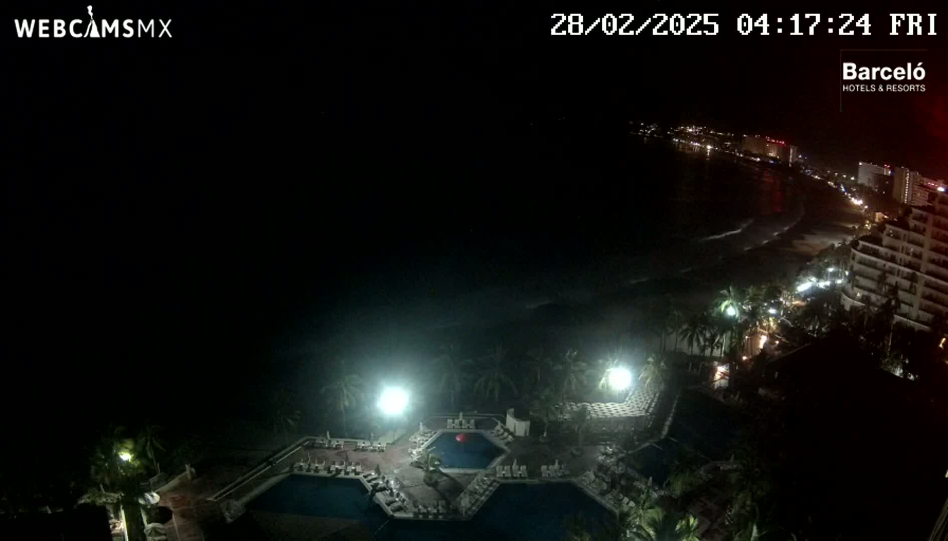 A nighttime, high-angle view shows a resort with multiple pools illuminated, overlooking a dark ocean with a faintly visible shoreline and city lights in the distance.