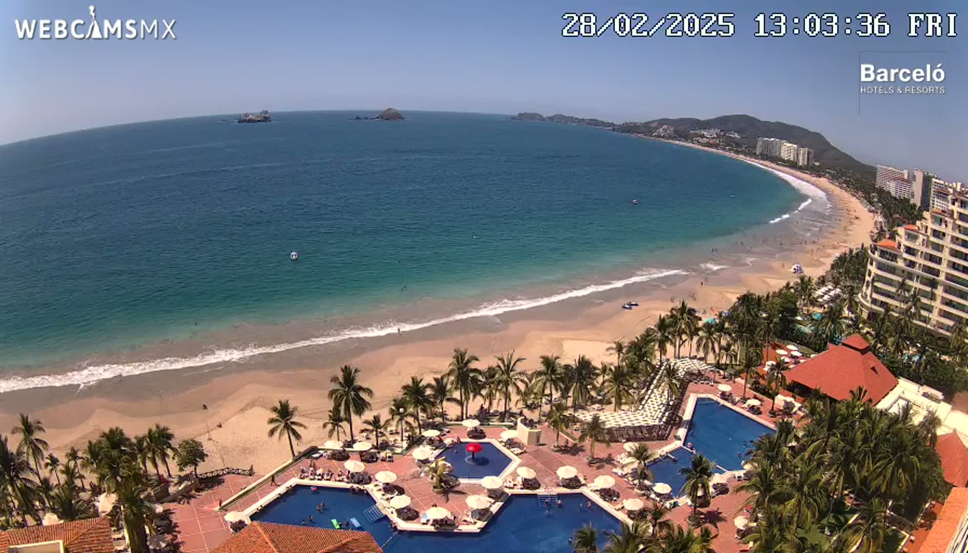 An aerial view shows a resort with multiple pools surrounded by palm trees next to a sandy beach and a calm ocean under a clear sky.