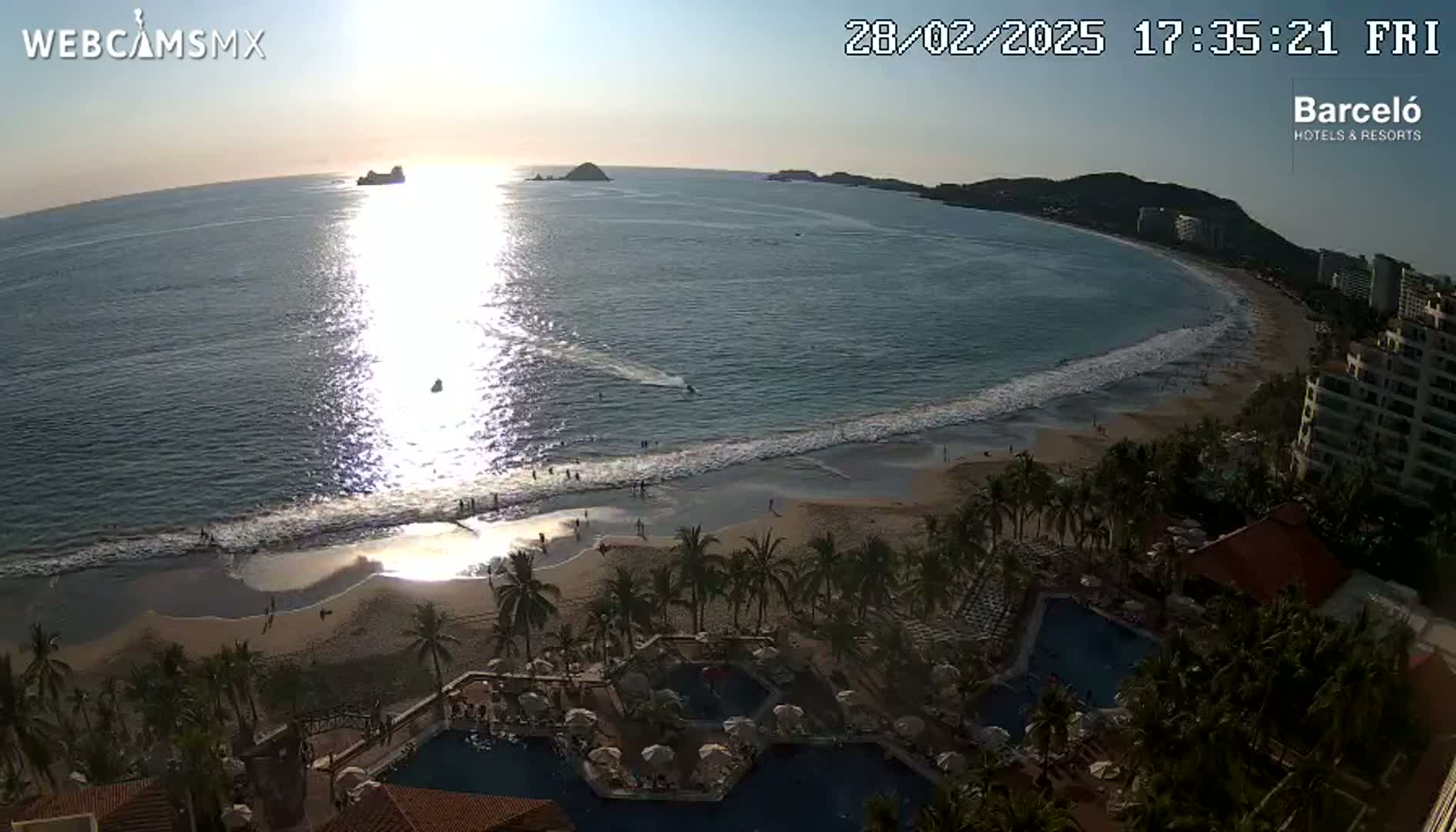 An ocean beach with calm waves at sunset is viewed from above, showing a resort with pools and palm trees in the foreground.