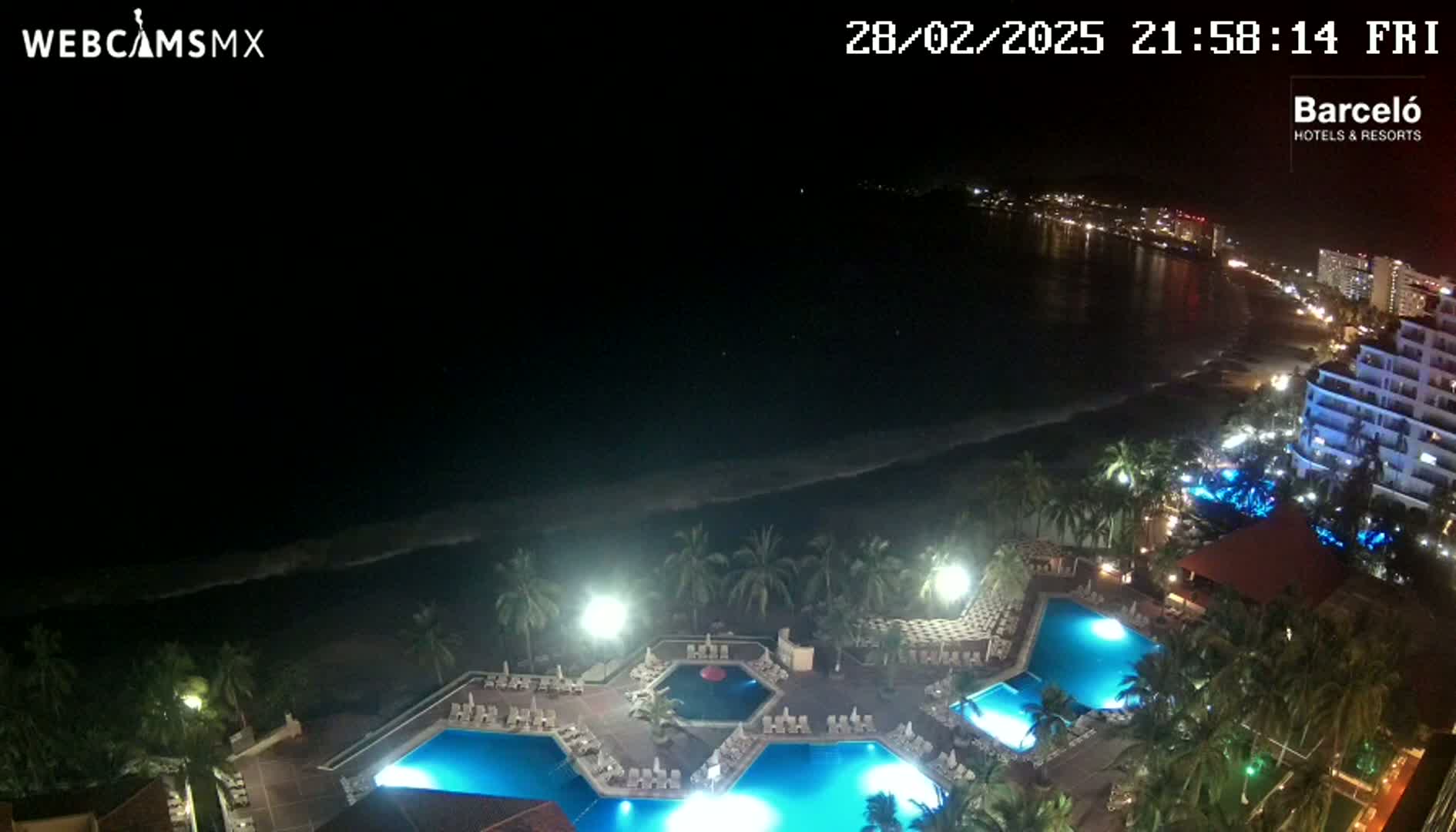 An aerial nighttime view shows a resort with illuminated swimming pools next to a dark ocean and a brightly lit beach.