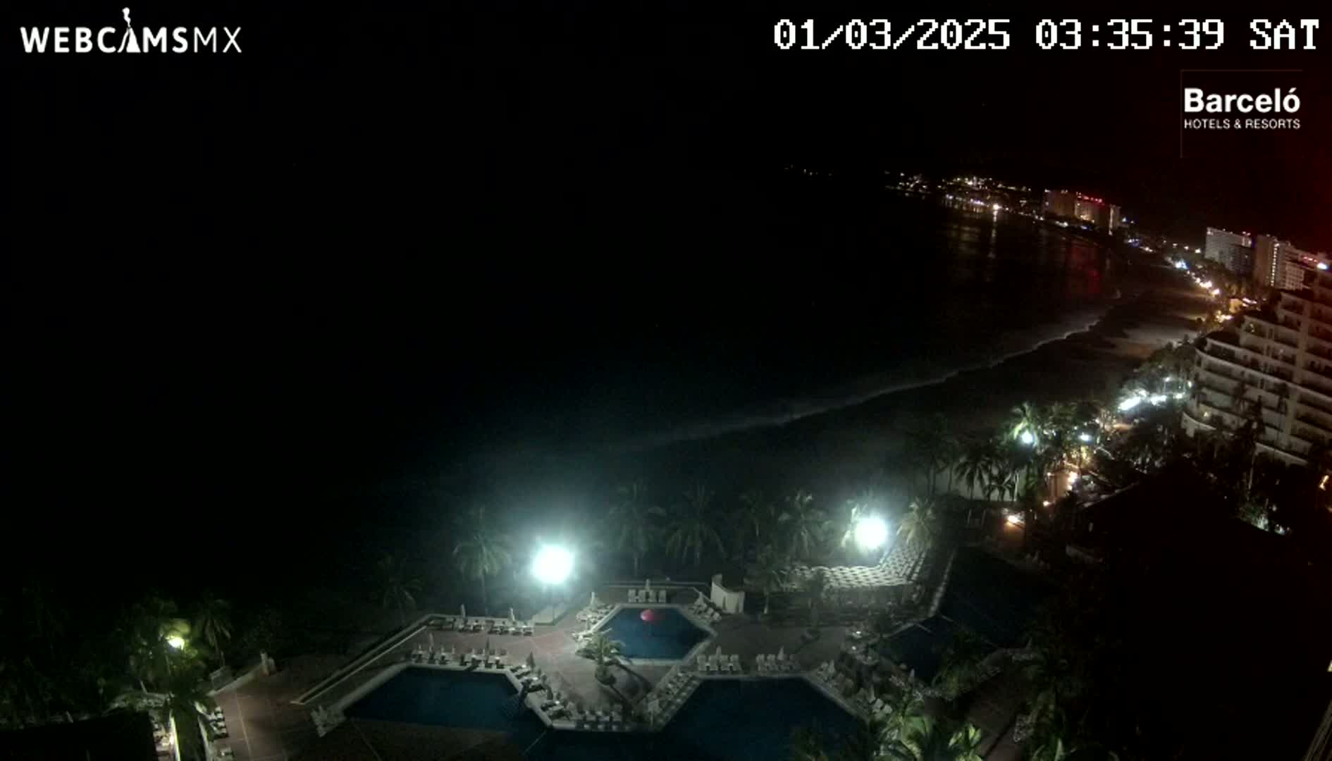 A nighttime view of a resort with multiple pools, surrounded by palm trees, overlooking a dark ocean and a dimly lit beach with buildings in the distance.